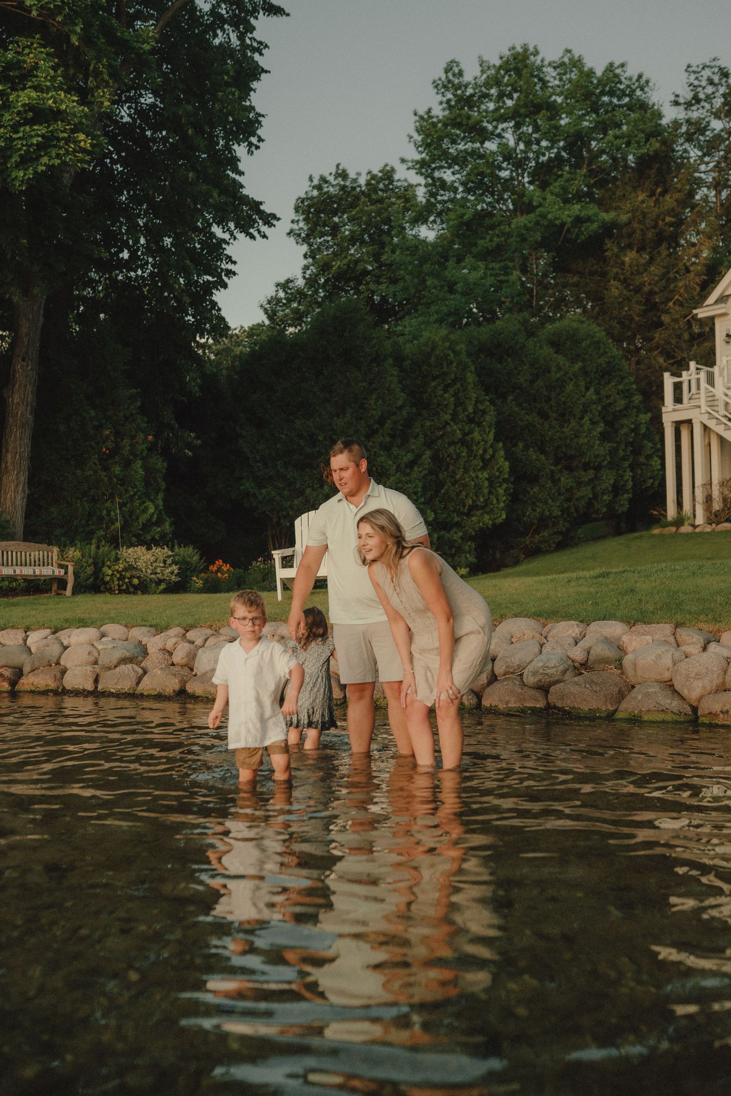 Family of four wades together in shallow lake water at sunset near a stone wall.