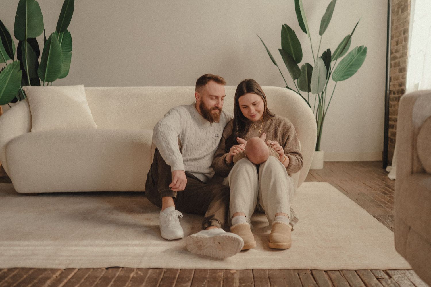 A couple relaxes together on a cozy beige carpet in a minimalist room with potted plants in the background.