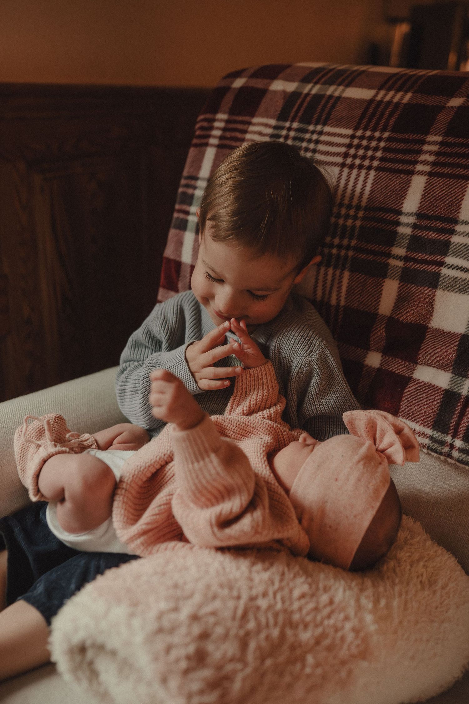 A child in a gray sweater cuddles and plays with a baby swaddled in pink on a plaid couch.
