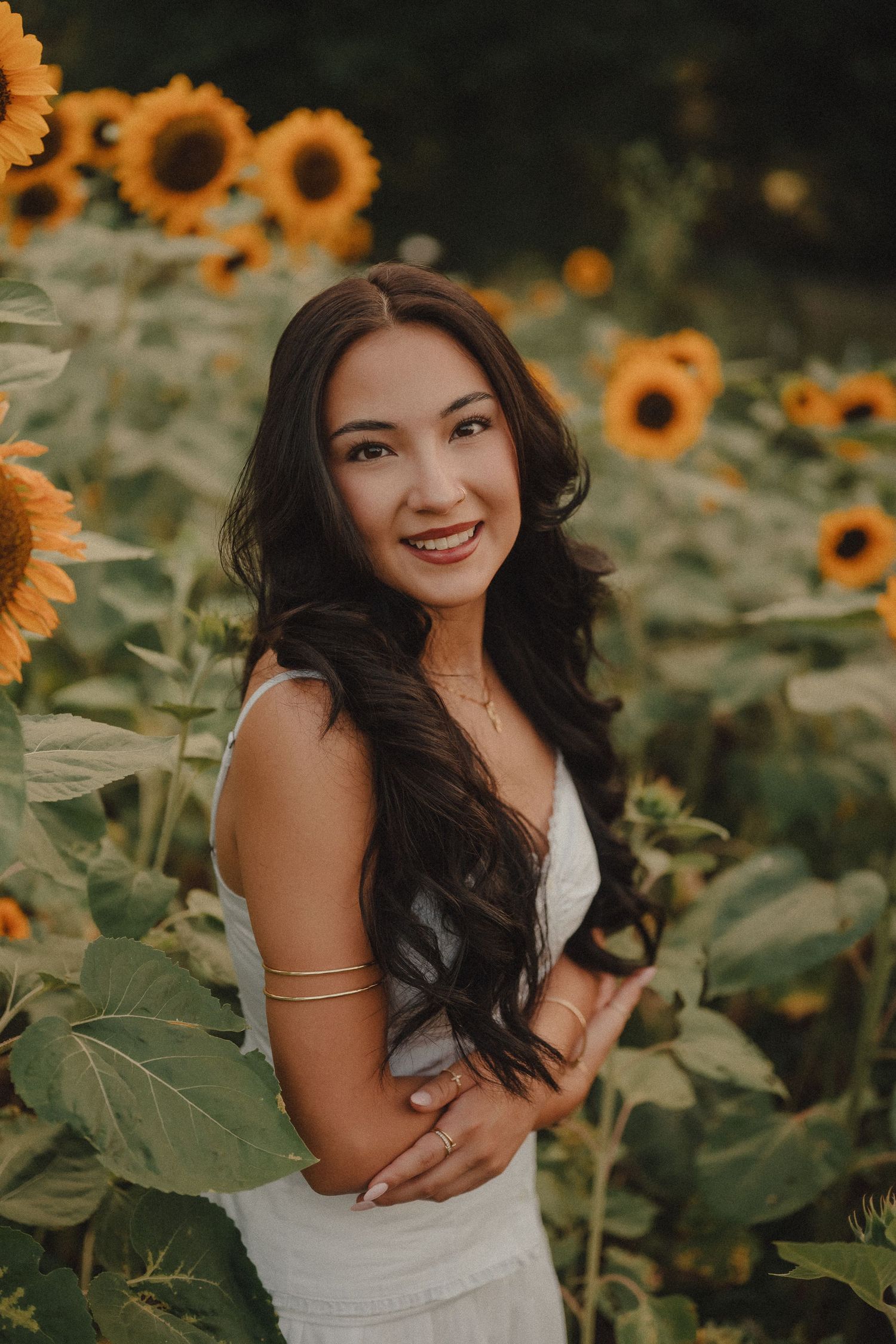 Smiling young woman in white dress posing among bright sunflowers in a sunflower field.