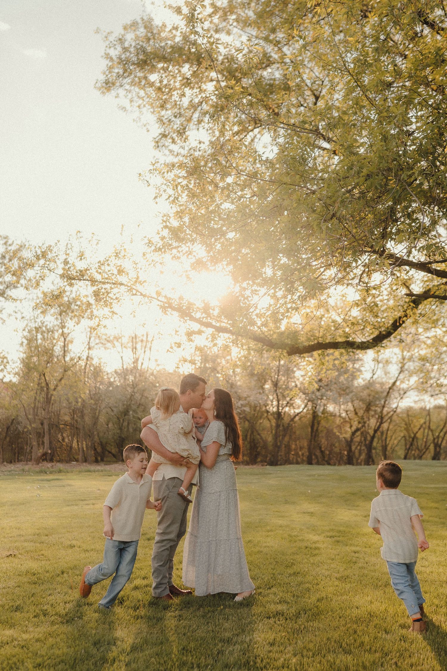 Family sharing a kiss at sunset in a lush green park with young children playing nearby.