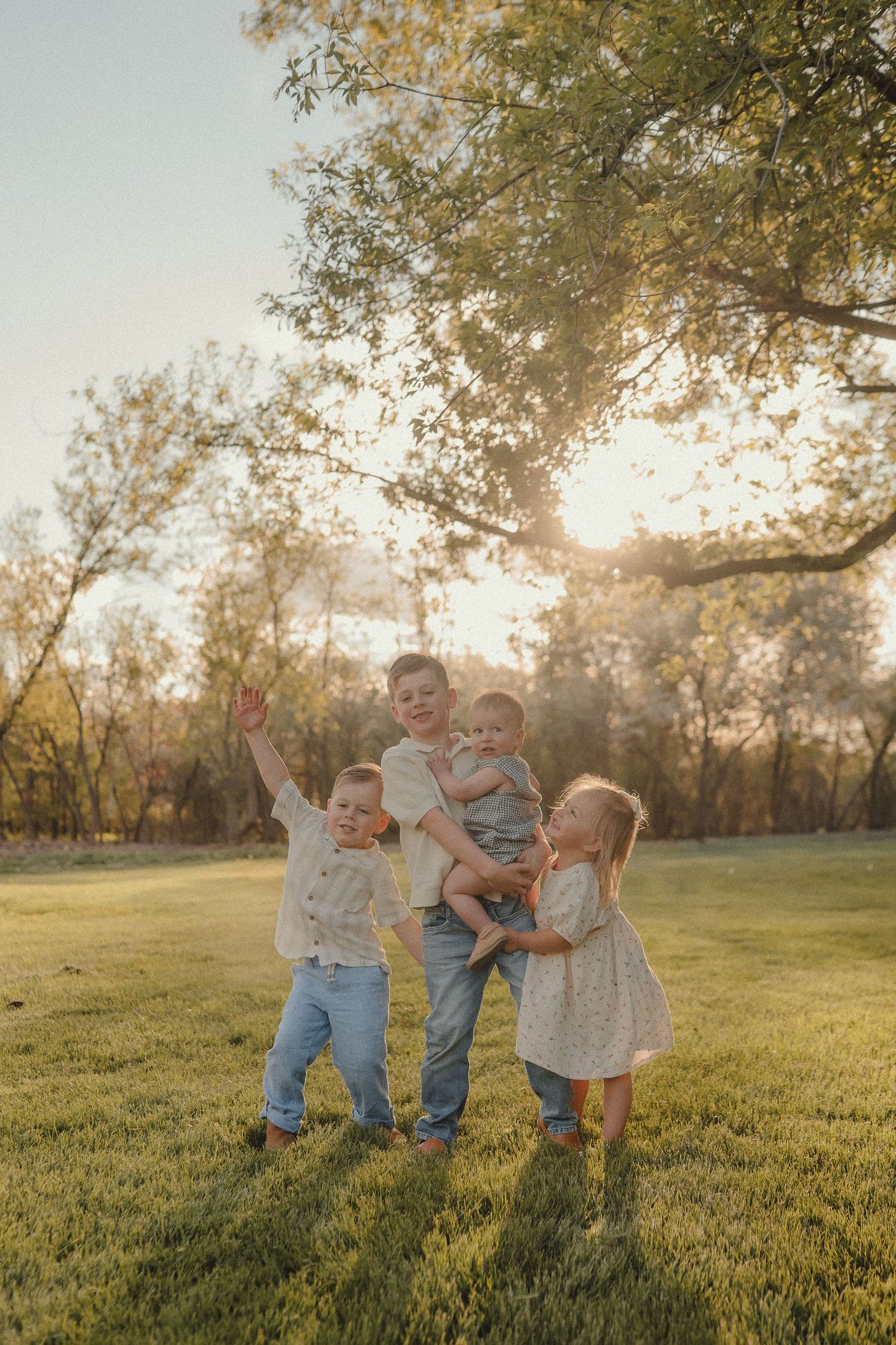 Father holding baby while two young children play around him in a sunlit green park.