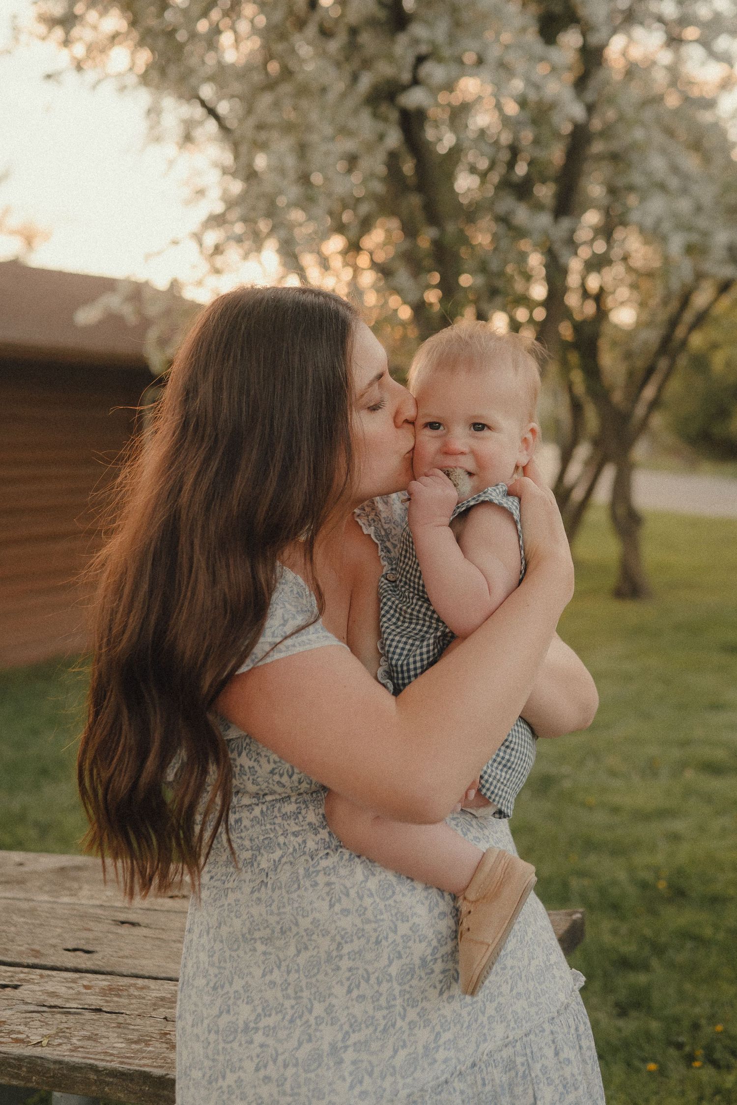 Loving mother kisses her happy baby girl while holding her near blooming trees at sunset outdoors.