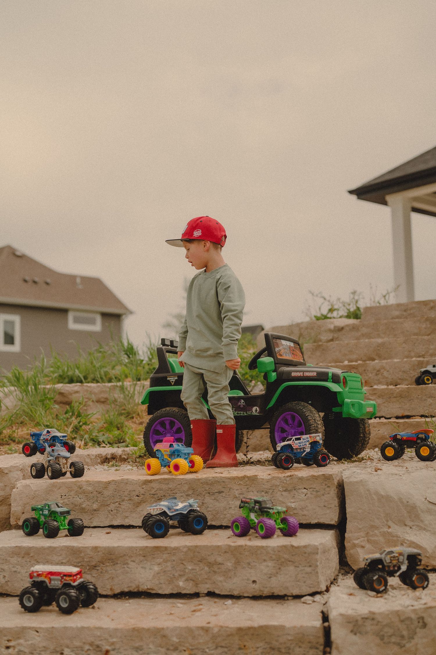 Young boy in red cap and boots stands on stone steps surrounded by colorful monster truck toys beside a ride-on jeep.