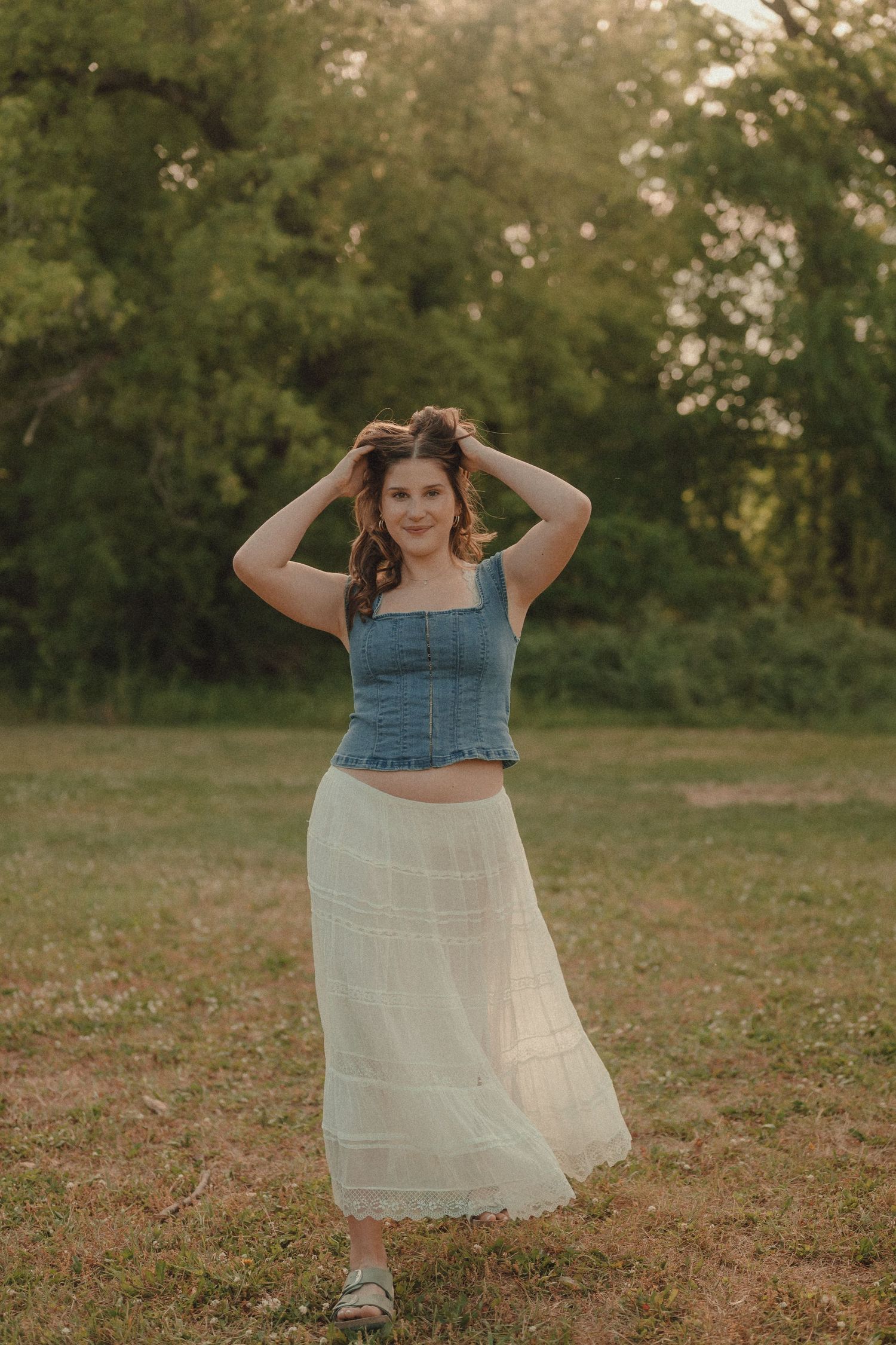 Young woman in denim crop top and white maxi skirt poses outdoors in a sunlit grassy field with green trees.