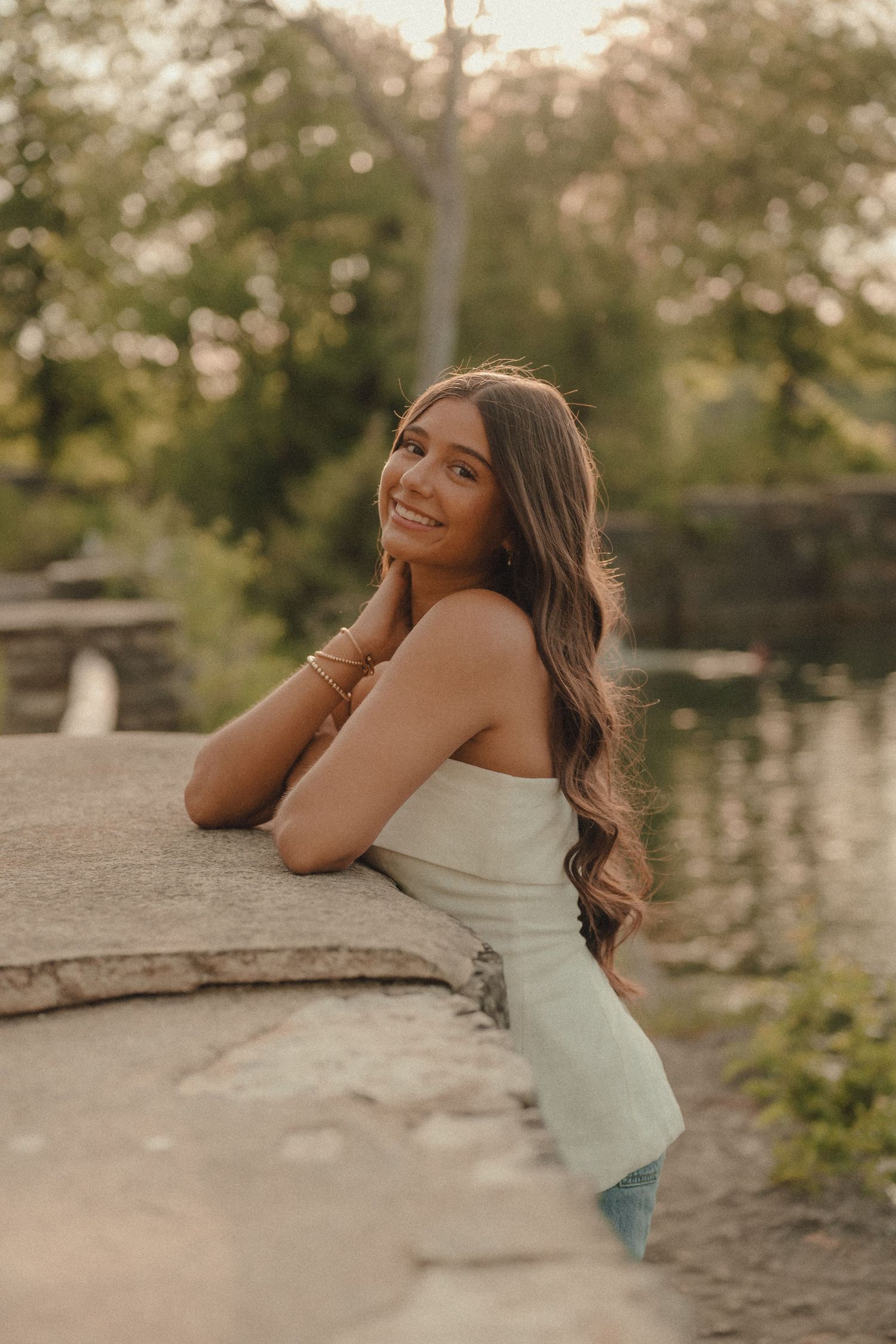 Smiling young woman in strapless dress leaning on stone wall near water at sunset.