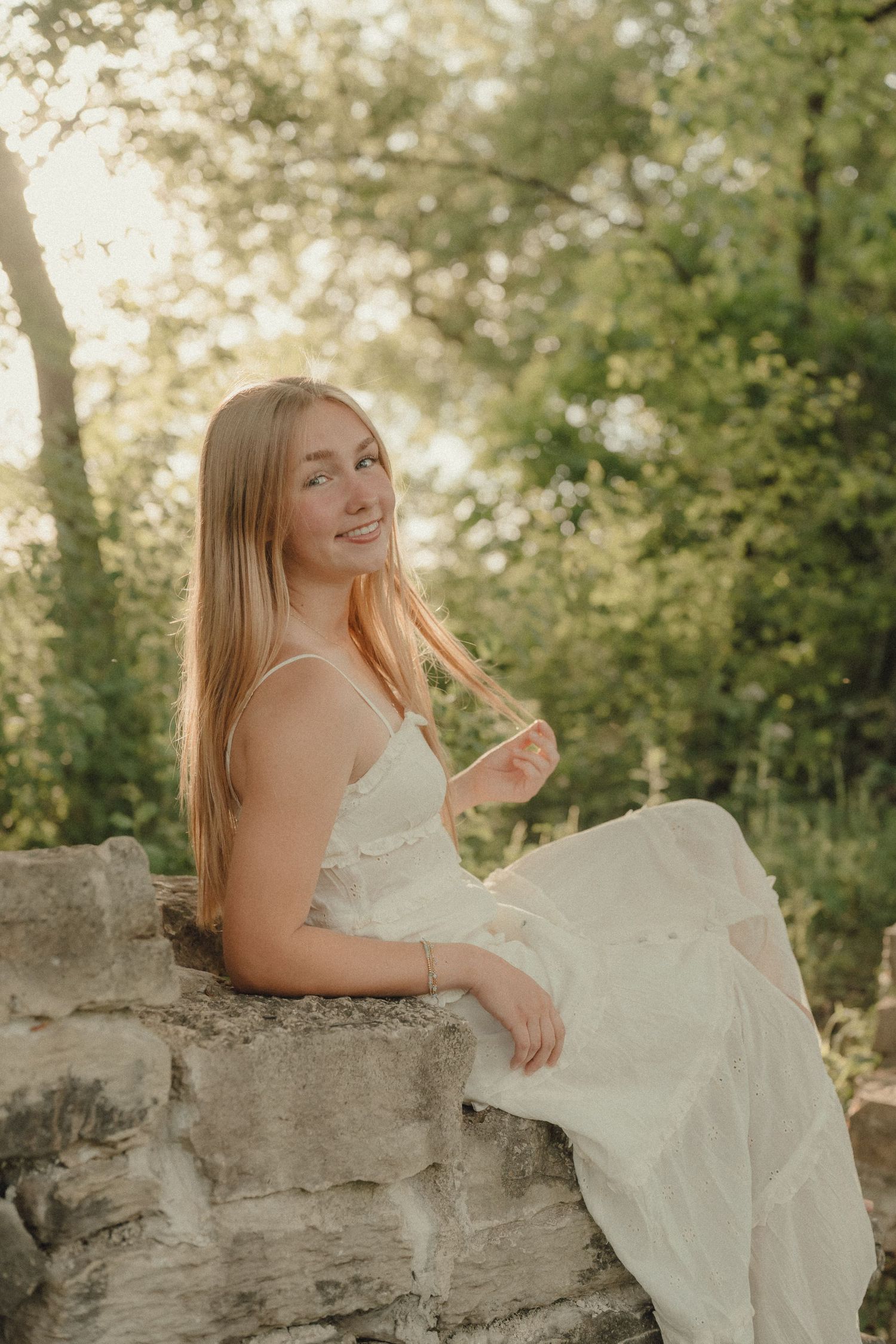 Young woman in white dress sitting on stone wall surrounded by lush green trees in golden hour light.