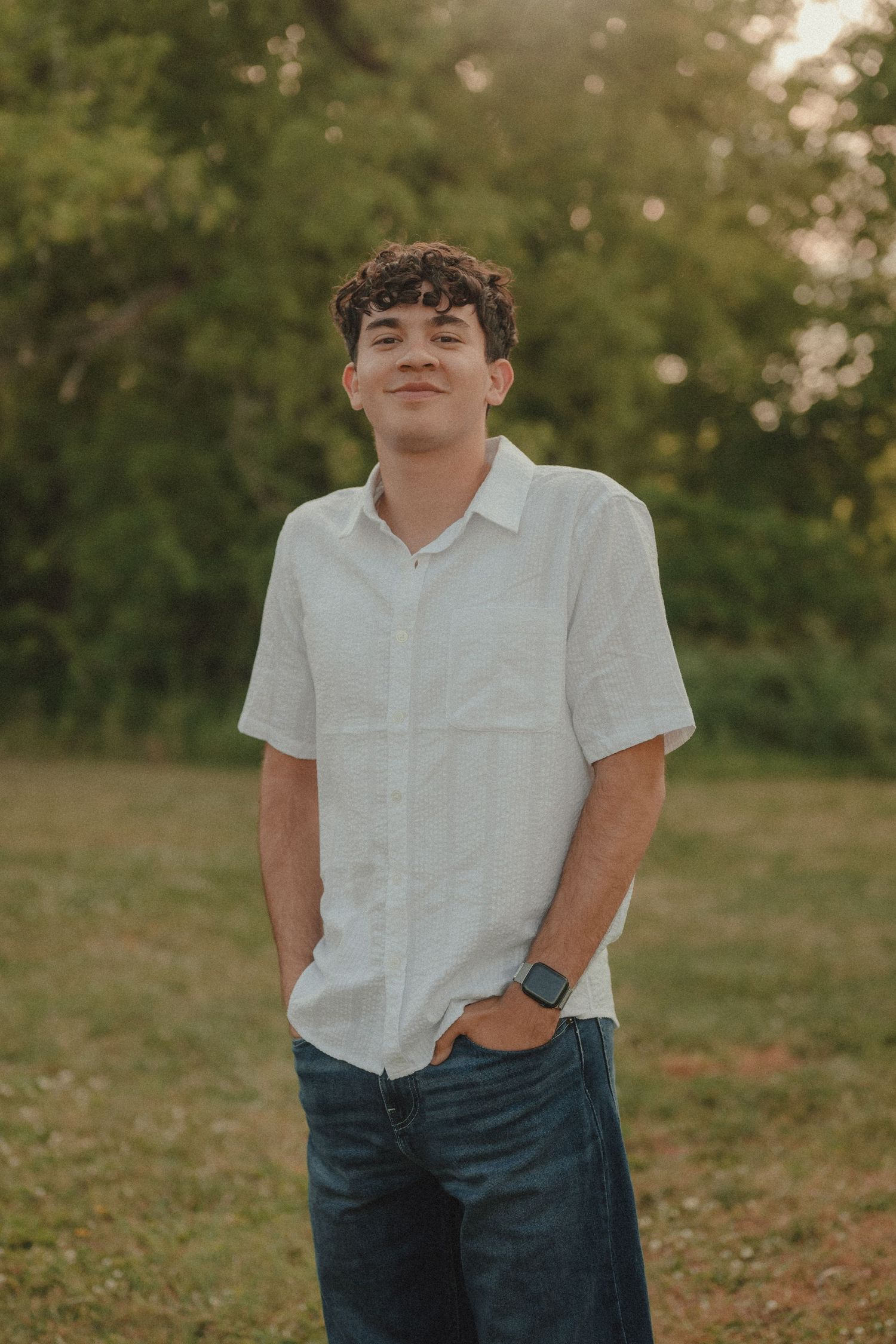 Young man in white button-up shirt and jeans standing outdoors in a green field during golden hour.