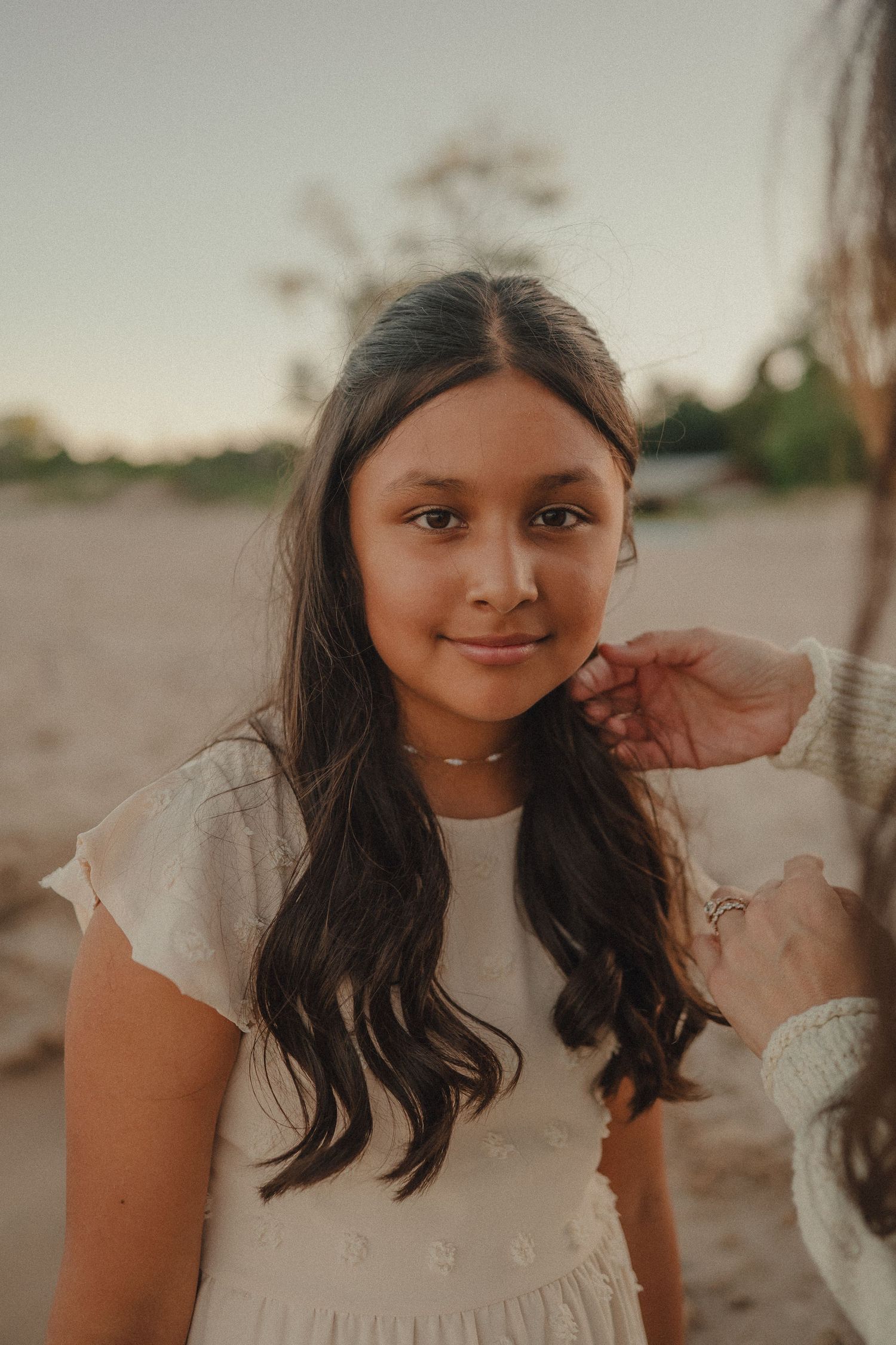 Young girl with long dark hair wearing white dress smiling softly at camera during beach sunset.