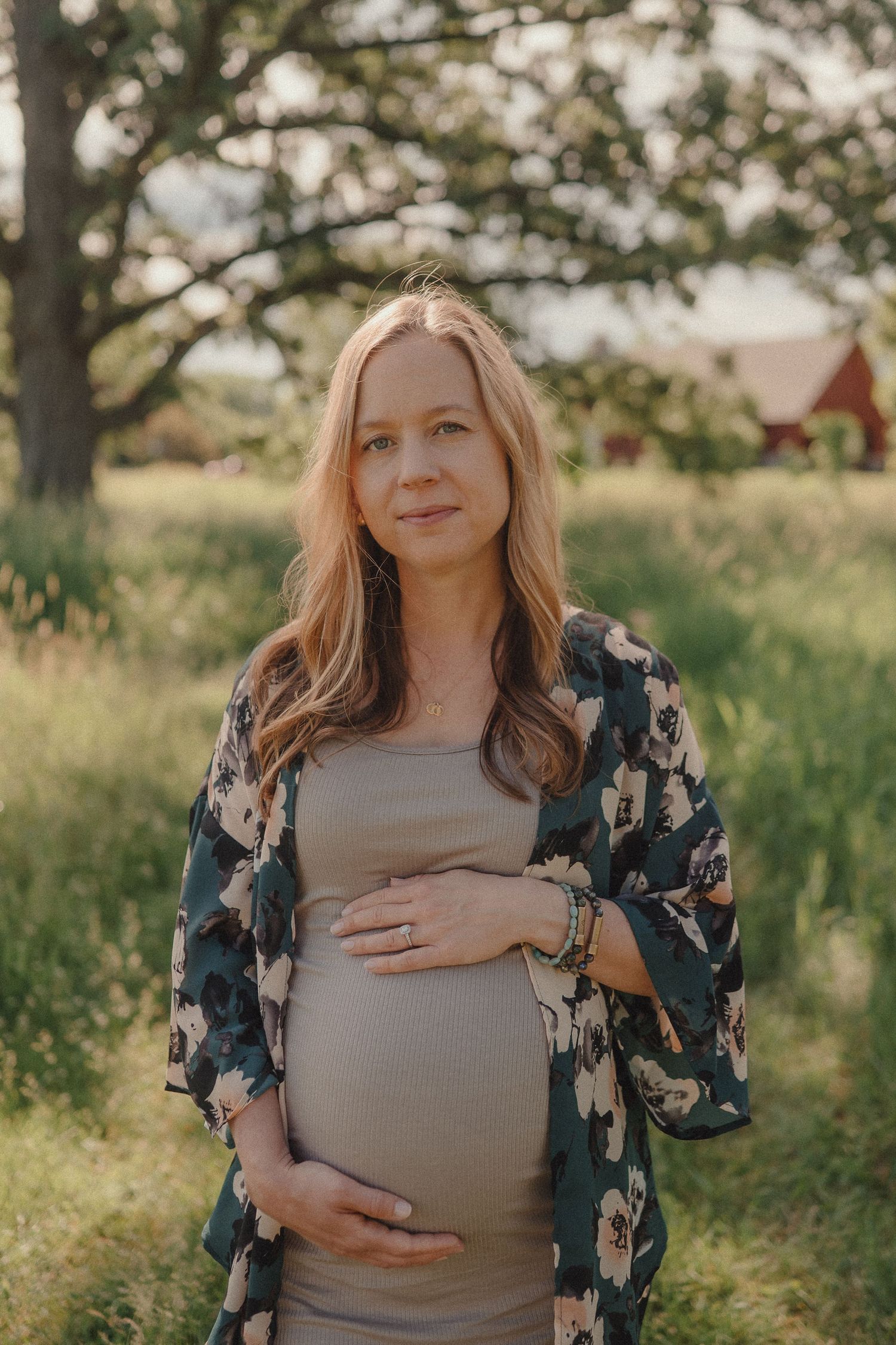 A pregnant woman in a floral kimono cradles her baby bump in a sunlit green field.