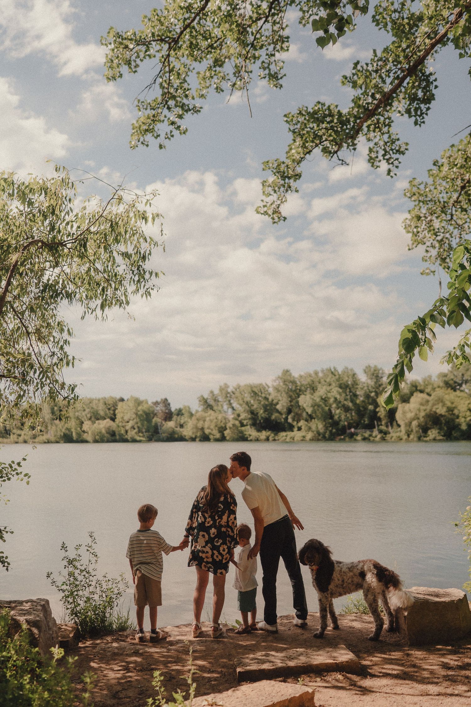 A family with two boys and a dog share a kiss by a scenic lake surrounded by lush green trees.