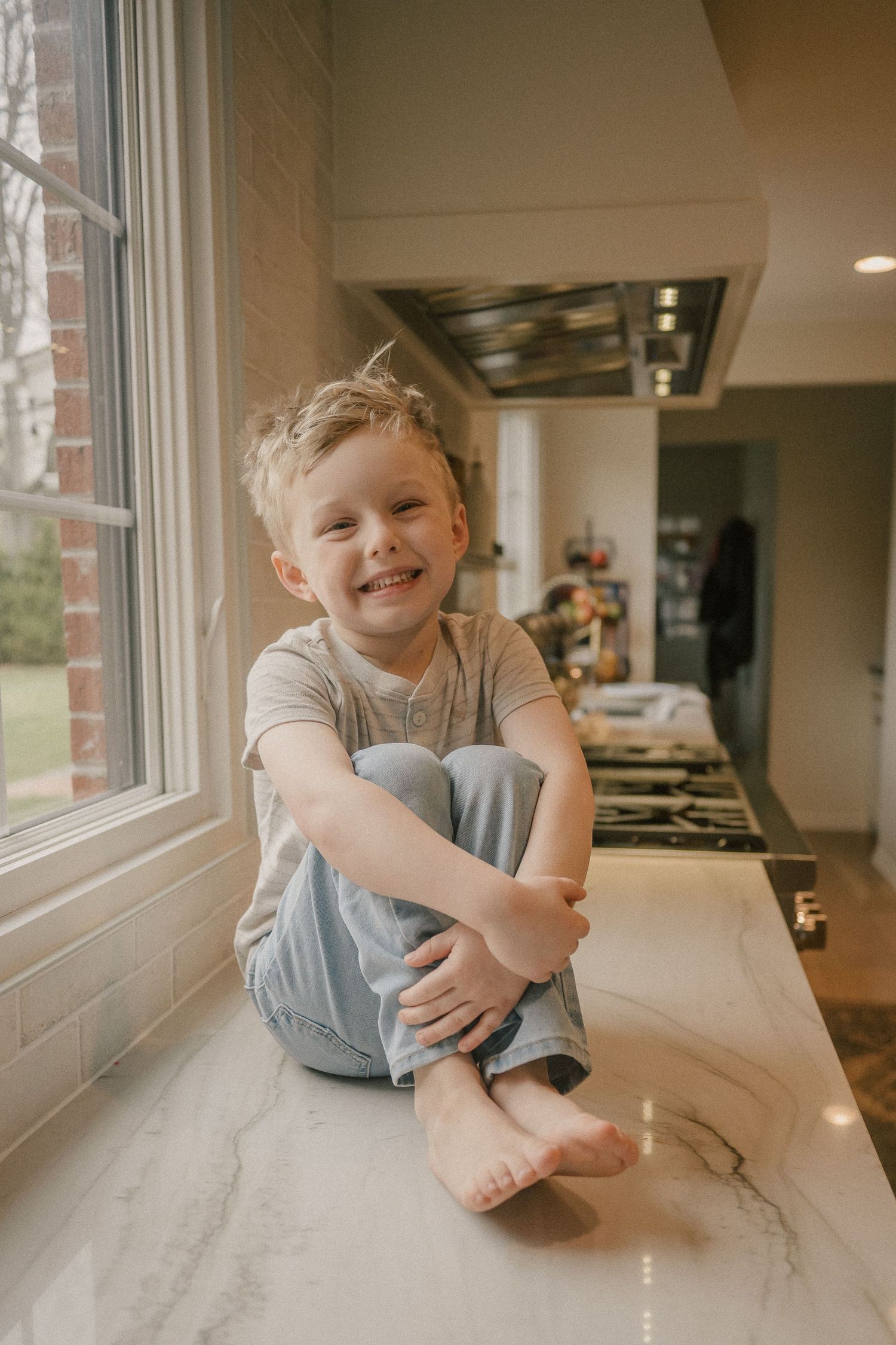 A happy young boy sits cross-legged on a marble kitchen counter smiling at the camera.