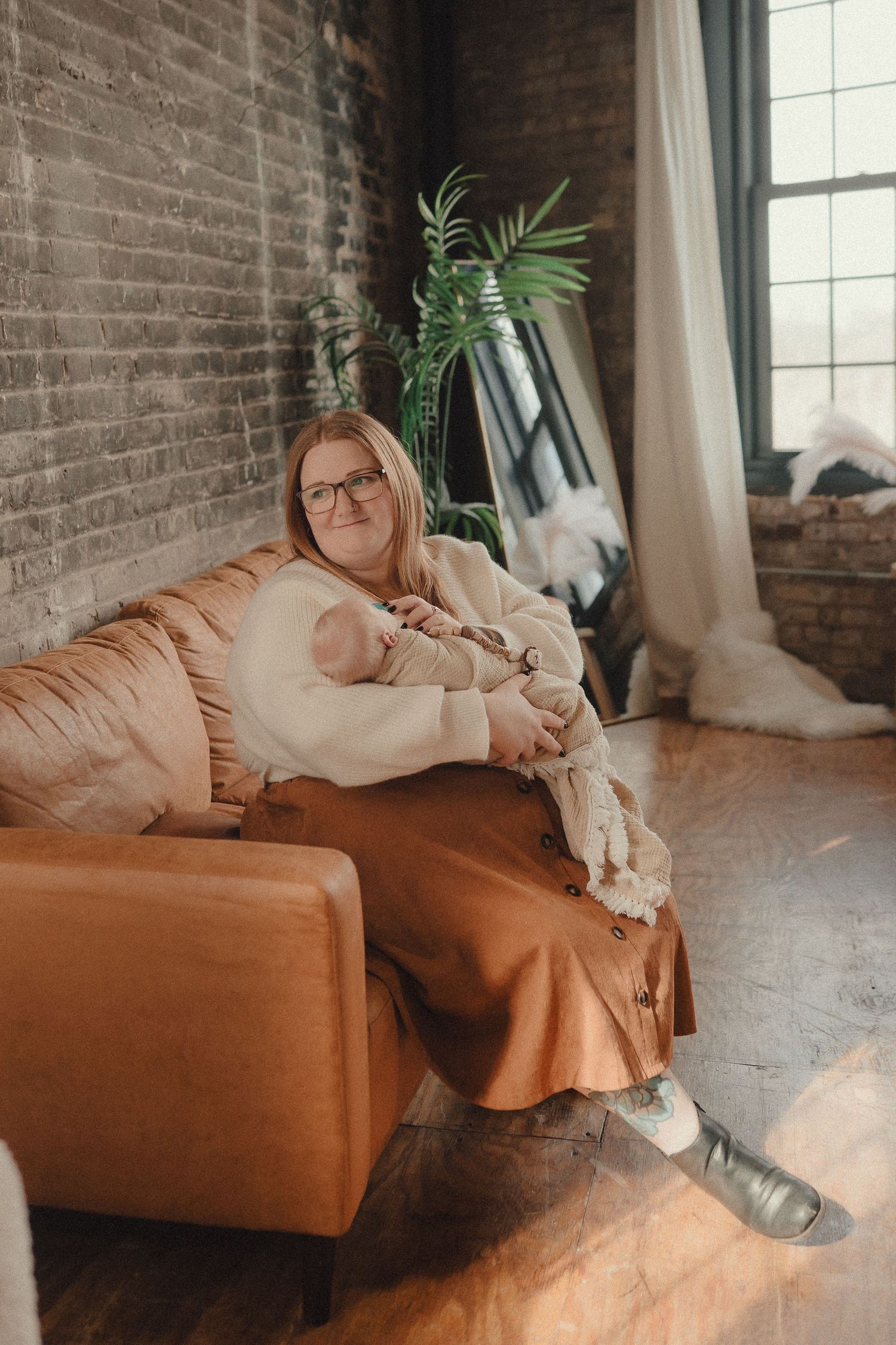 Woman in cream sweater and rust skirt sits relaxed on brown leather sofa in industrial loft with brick walls.