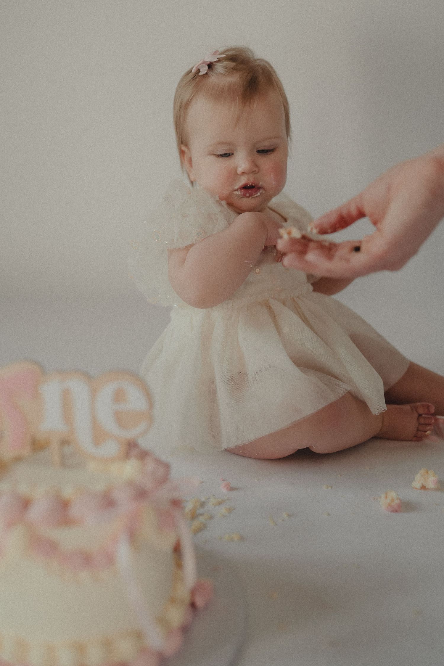 Baby girl in ivory dress tastes her first birthday smash cake with help during milestone photo session.