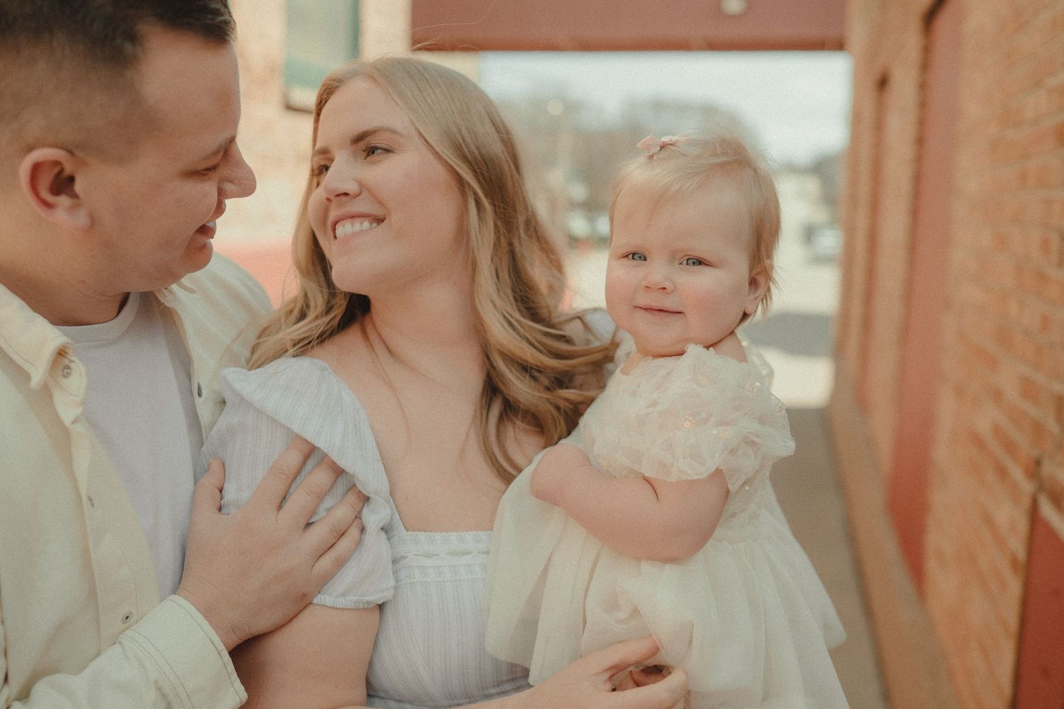 A smiling mother holds her baby girl while father looks on tenderly in warm outdoor light.