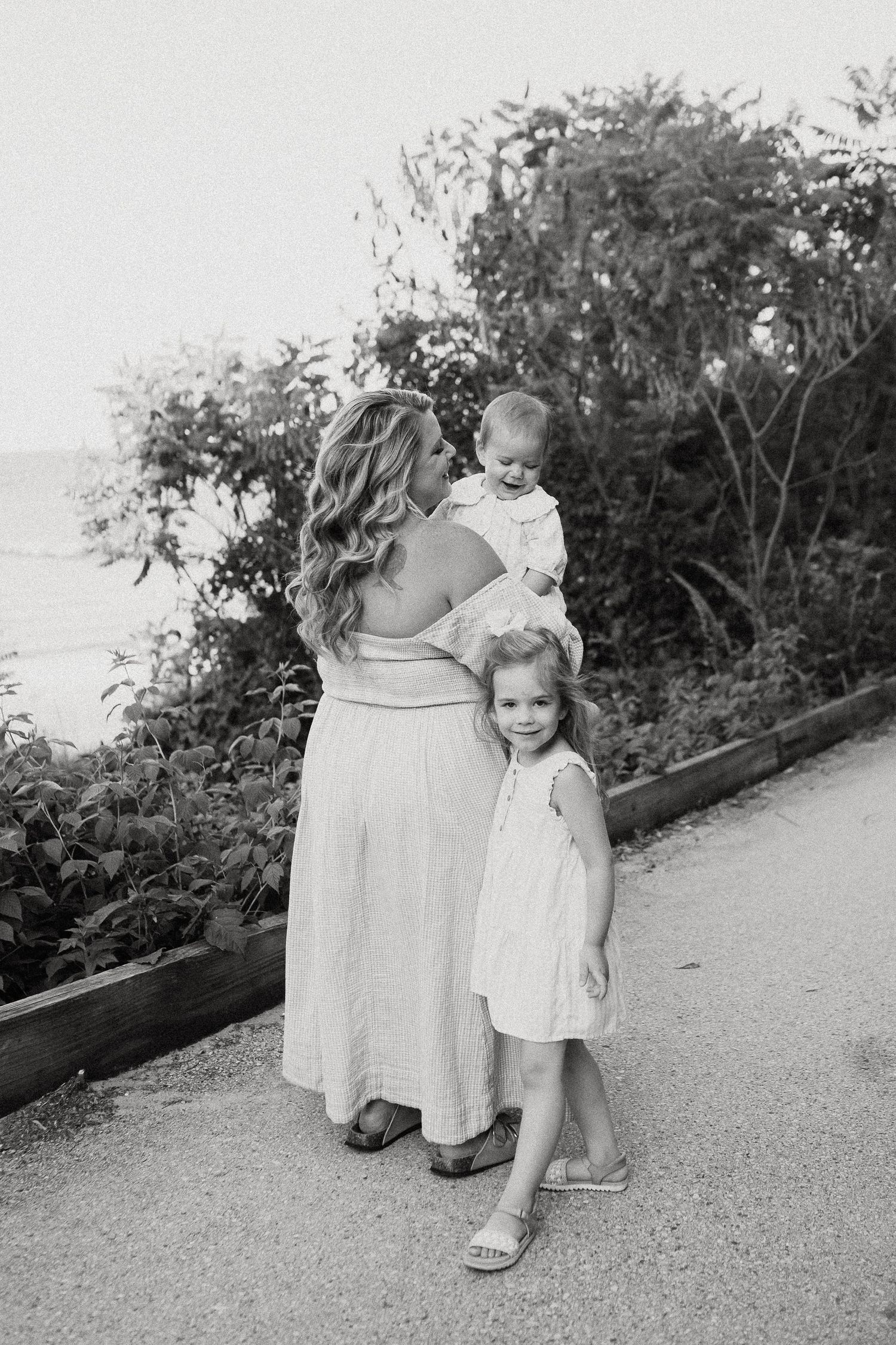 Mother holding baby while young girl hugs her, black and white family photo near lush greenery and ocean.