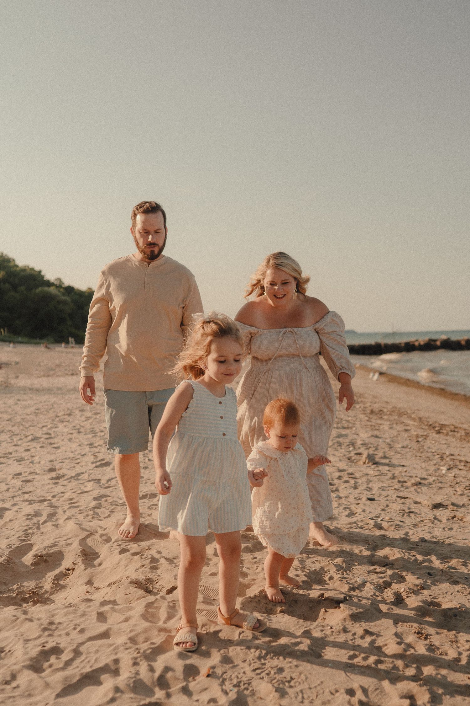 Family of four walking on sandy beach at golden hour, pregnant mother in flowing dress with two young daughters.
