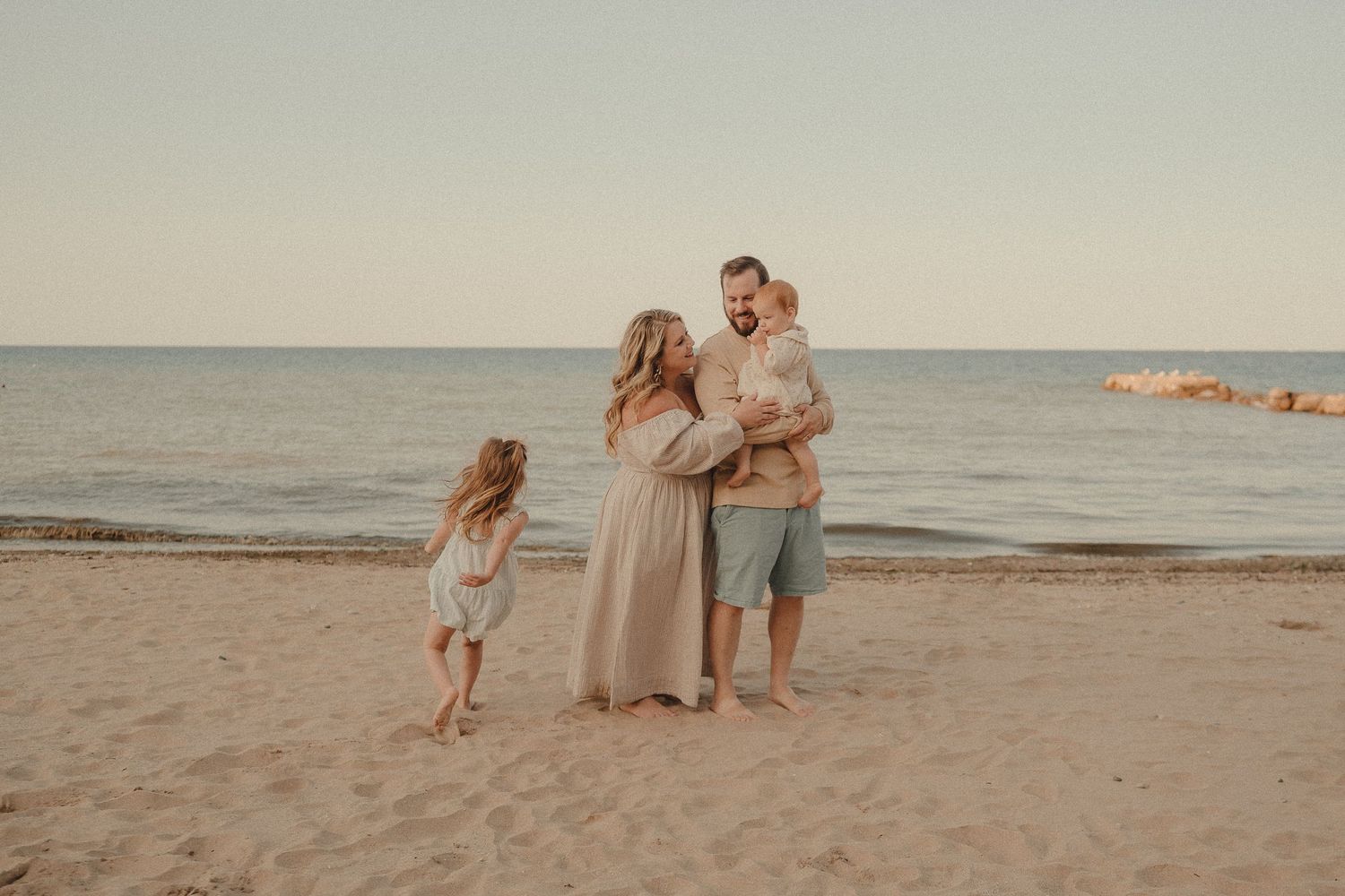 Happy family of four standing on sandy beach at sunset, parents holding toddler while daughter runs nearby.