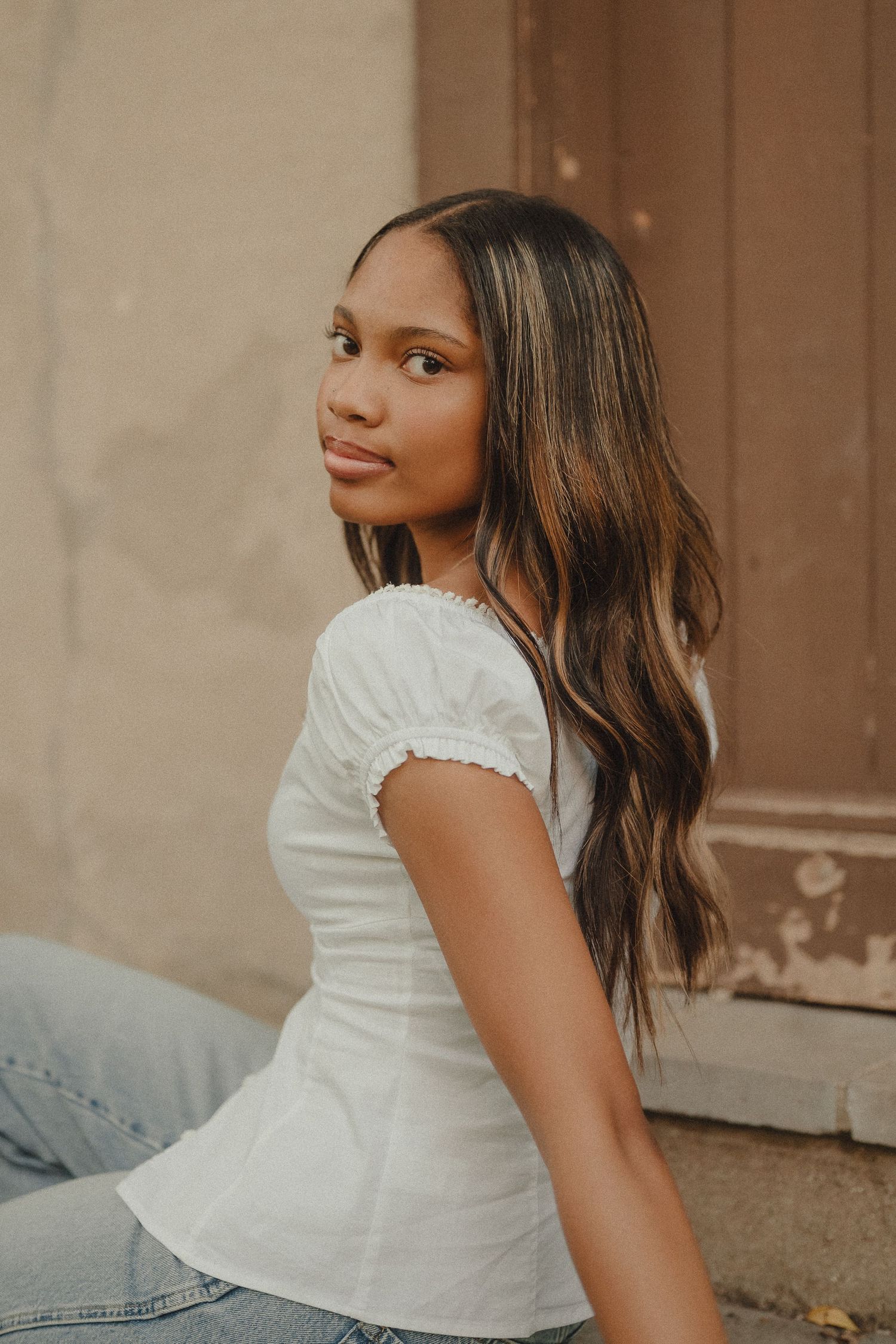Young woman with long wavy hair sits outdoors in a white smocked top and jeans, glancing over her shoulder.