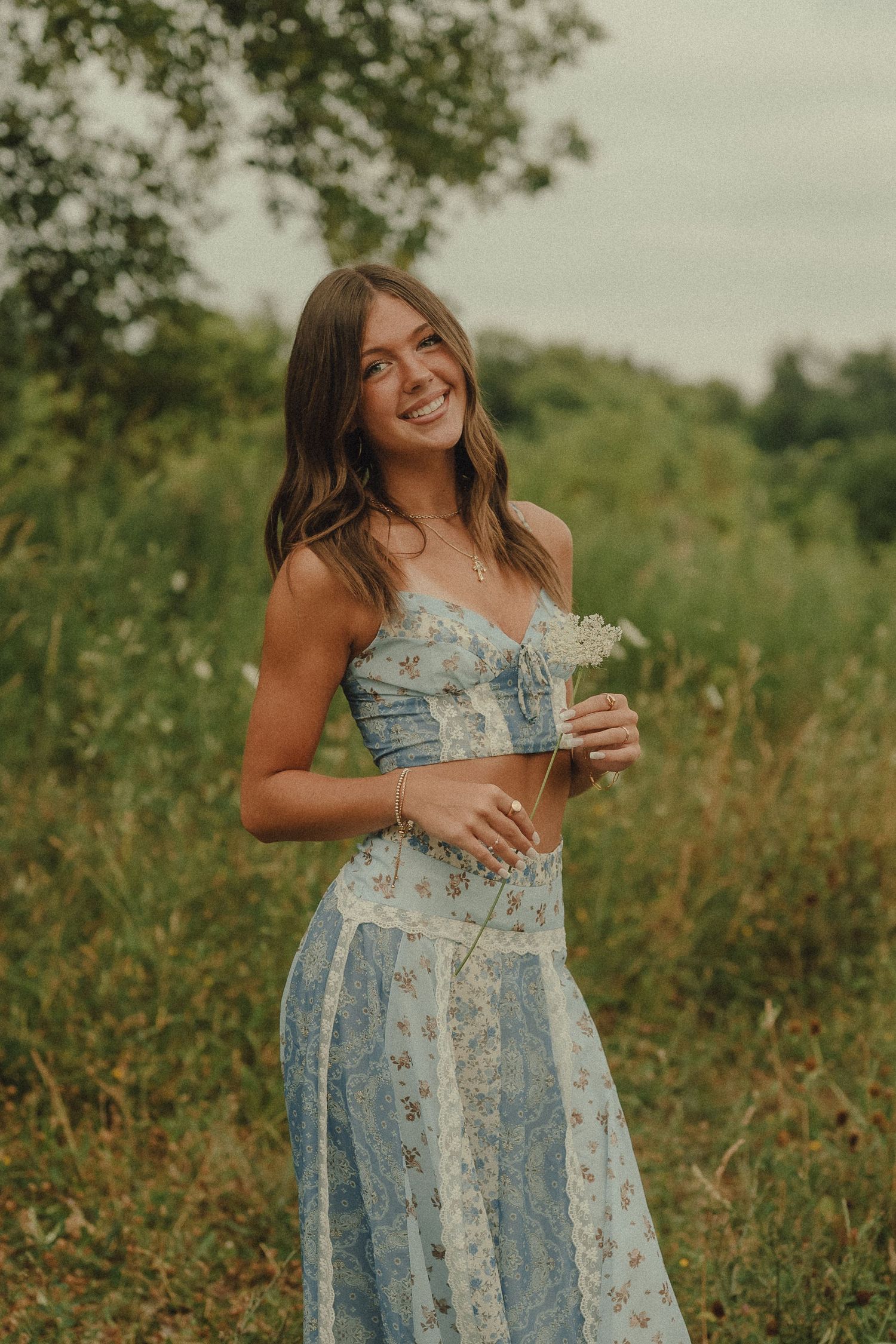 Smiling young woman in blue floral crop top and maxi skirt holding wildflowers in a lush green field.