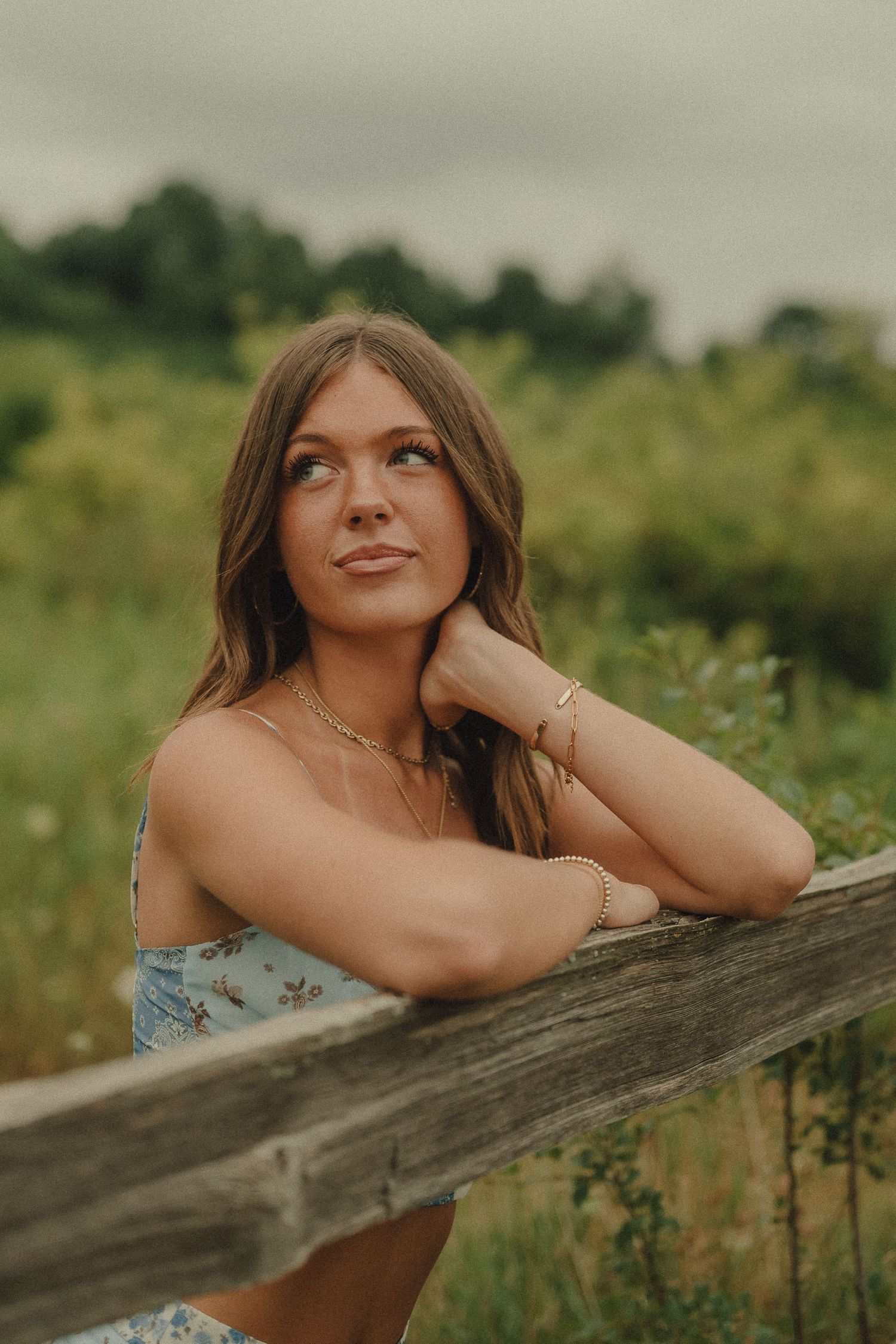 Young woman in blue floral dress leaning on a rustic wooden fence gazing upward in a green meadow.