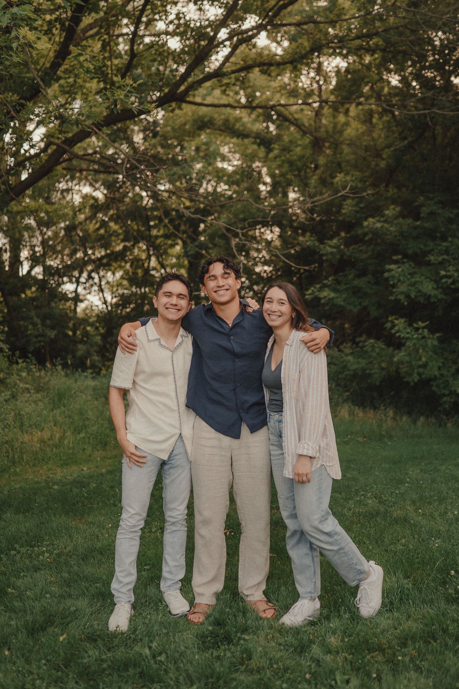 Three young adults smiling and posing together outdoors in a lush green park at golden hour.
