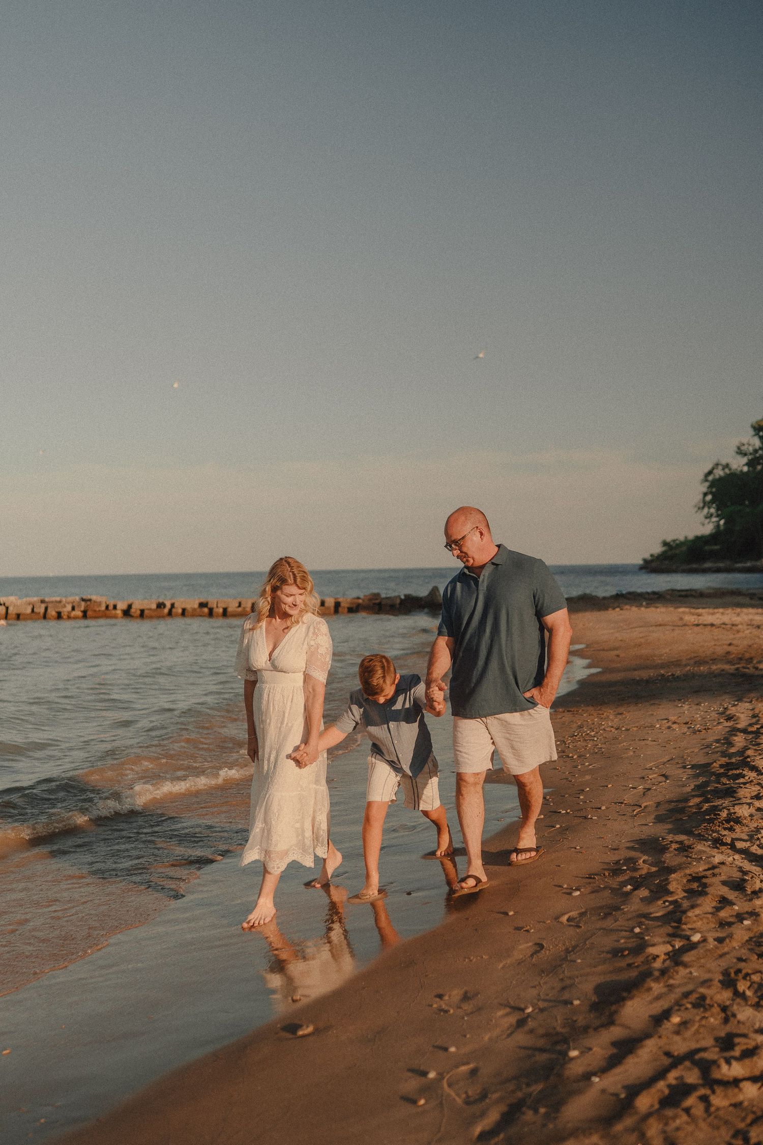 Family of three walking hand in hand along the shoreline at golden hour sunset.
