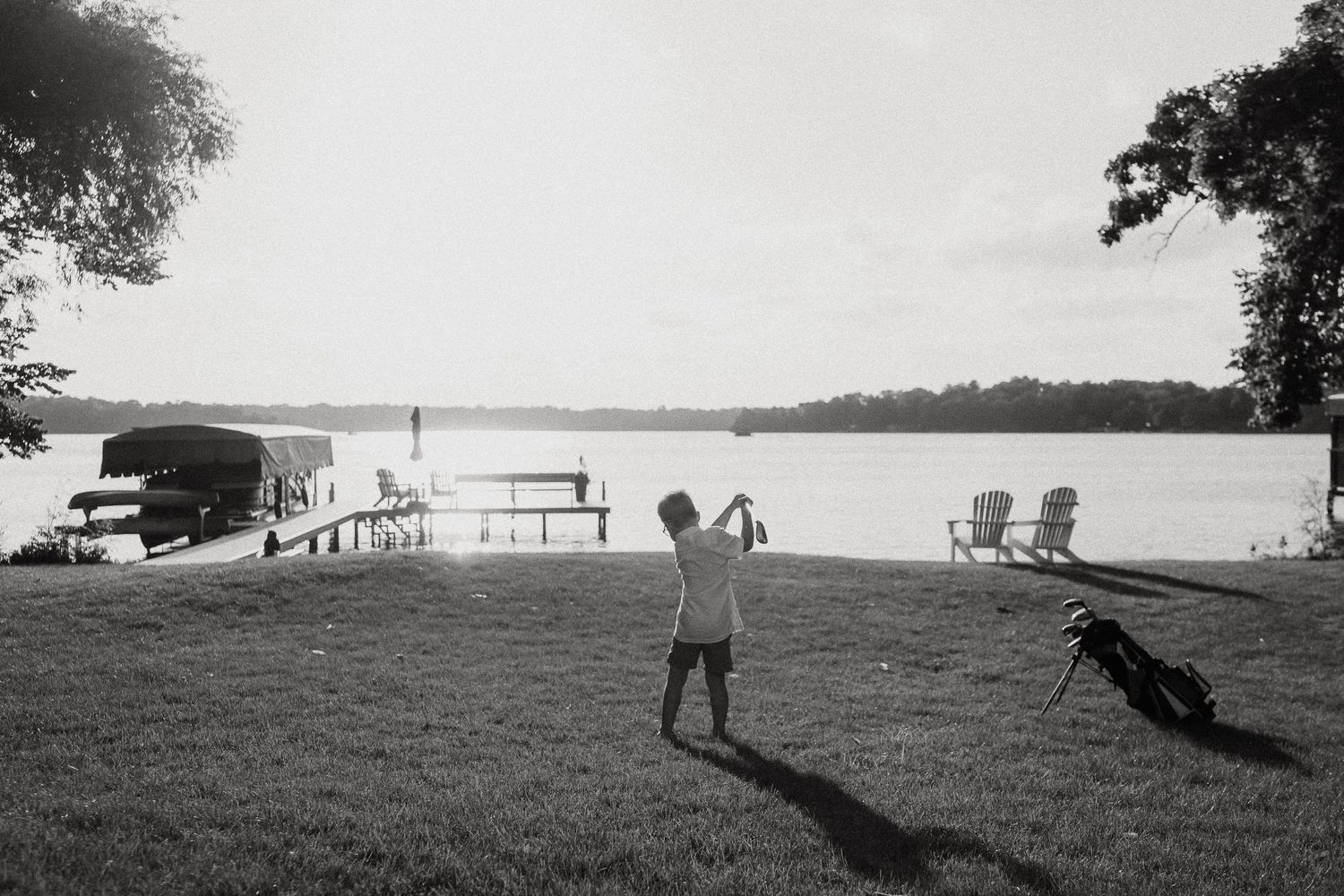 Child swinging a golf club on a lakeside lawn with Adirondack chairs and a dock in the background.
