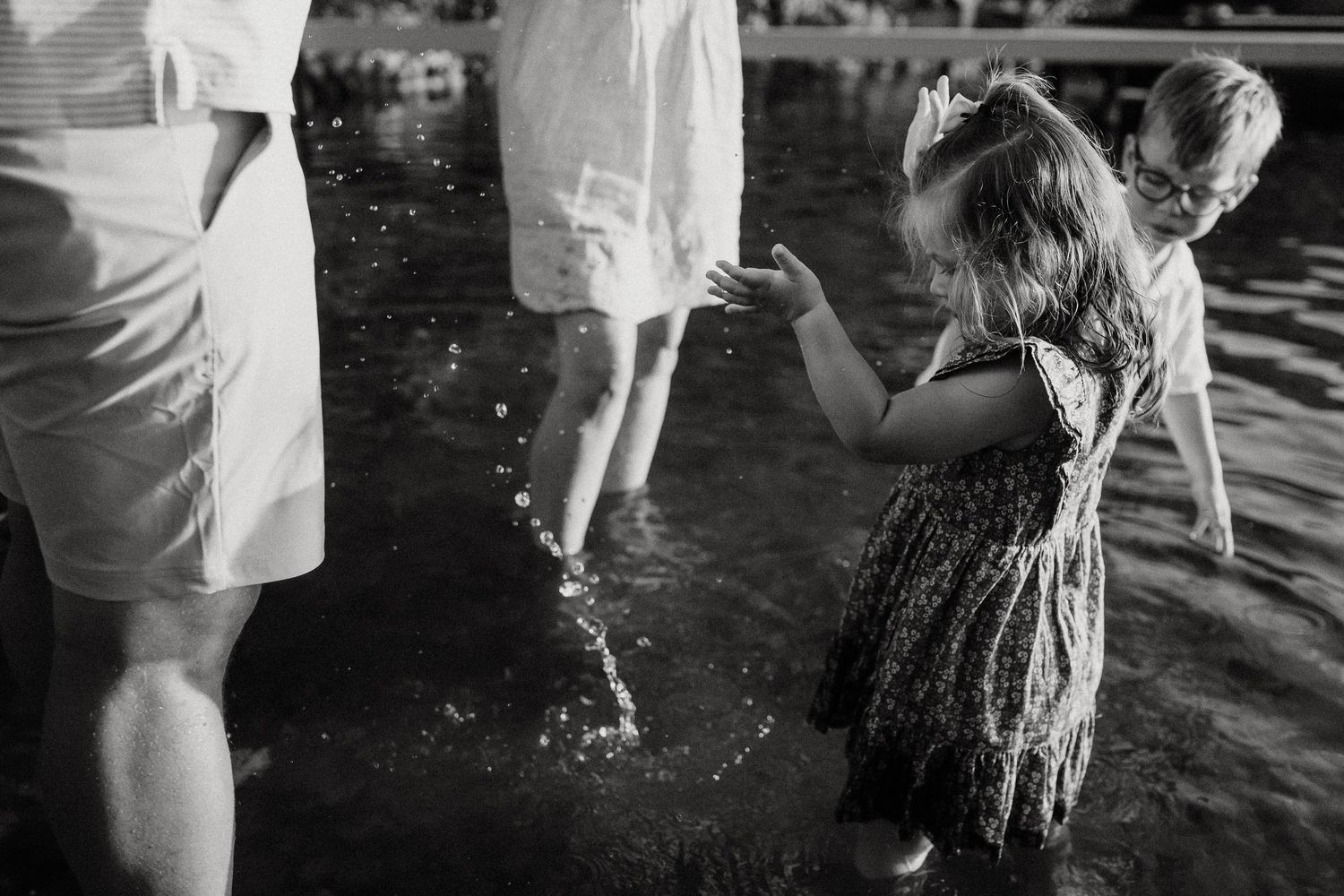 Black and white photo of a little girl splashing water at a lakeside with adults nearby.
