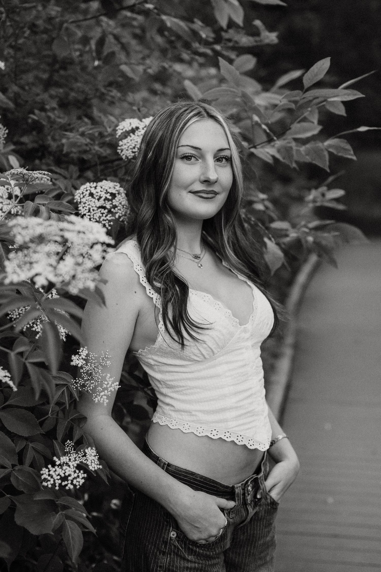 Young woman in white lace crop top and jeans poses beside blooming flowers in black and white portrait.
