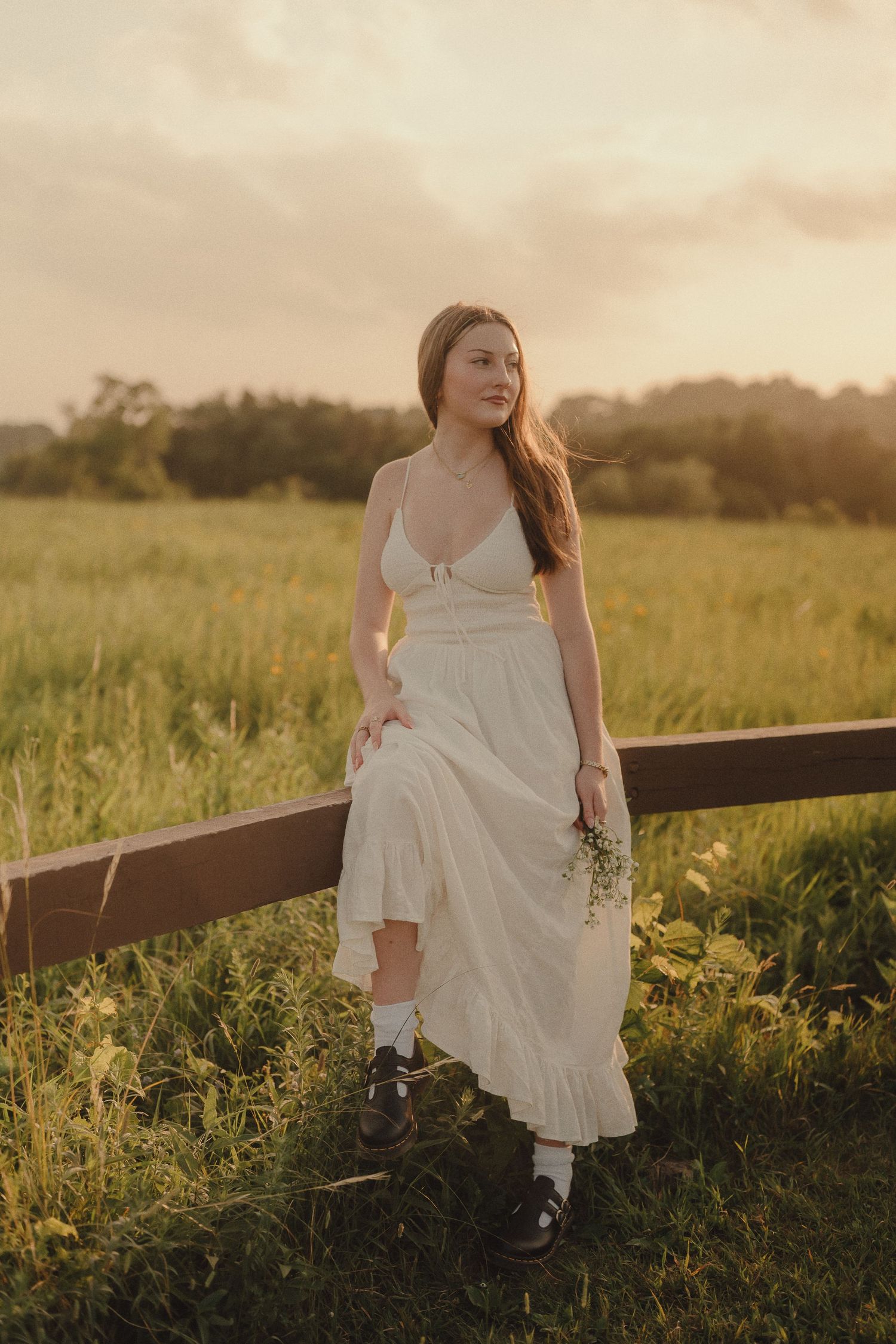Young woman in white dress holding wildflowers, leaning on a fence in a golden sunset field.