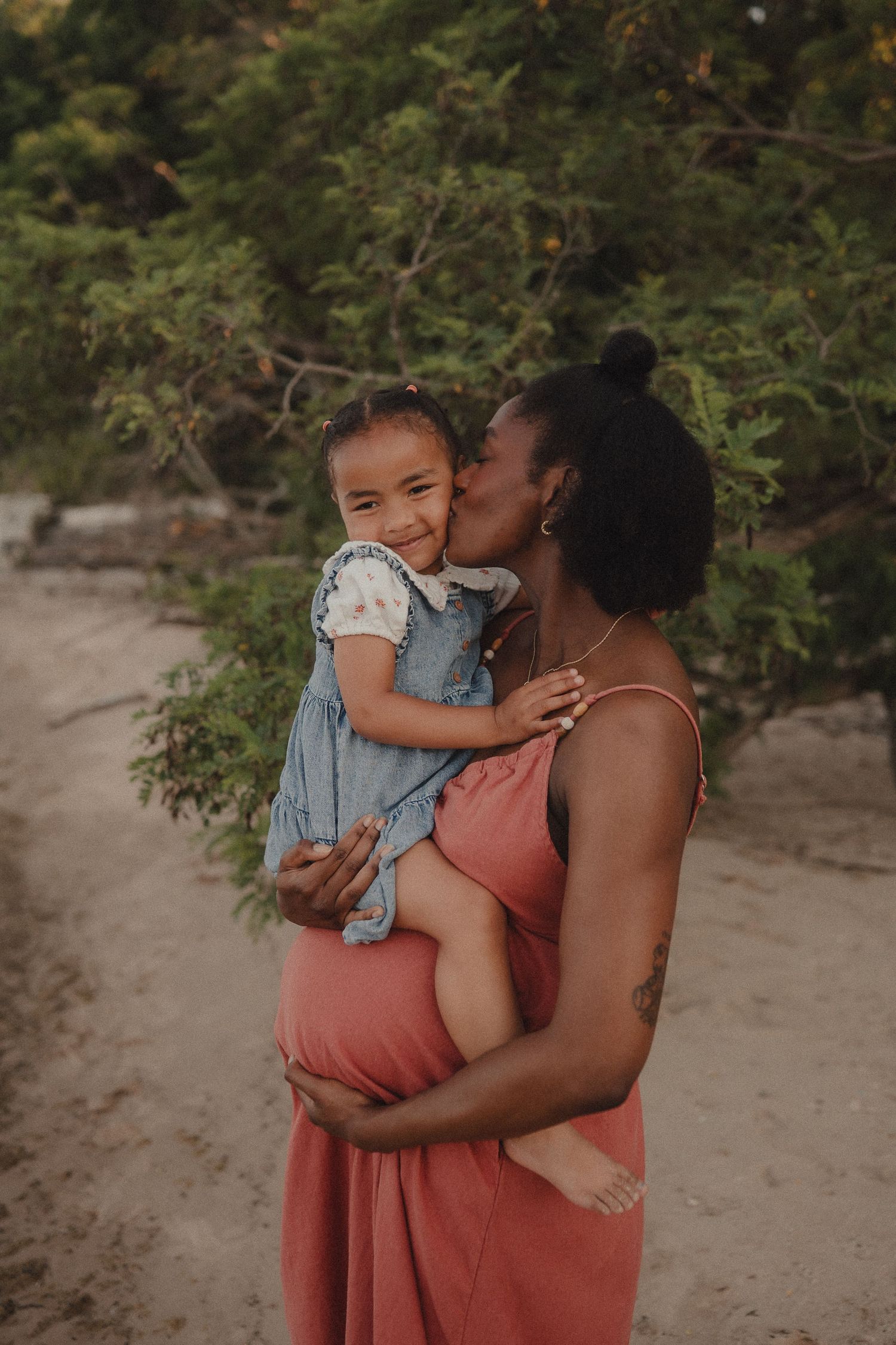 A pregnant Black woman in a coral dress kisses her smiling toddler daughter on a sandy beach.
