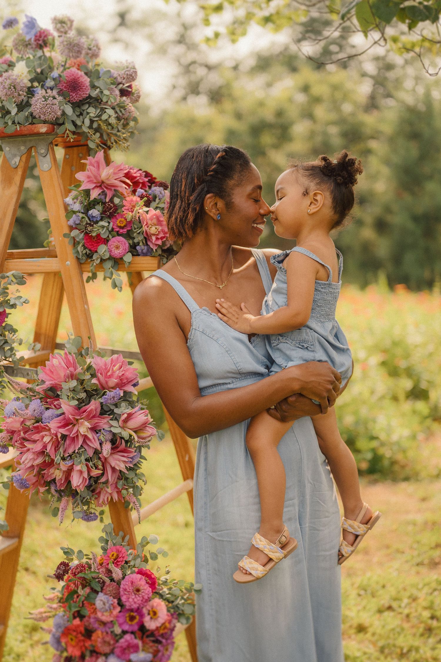Two people in light blue dresses share a tender moment among pink floral arrangements outdoors.