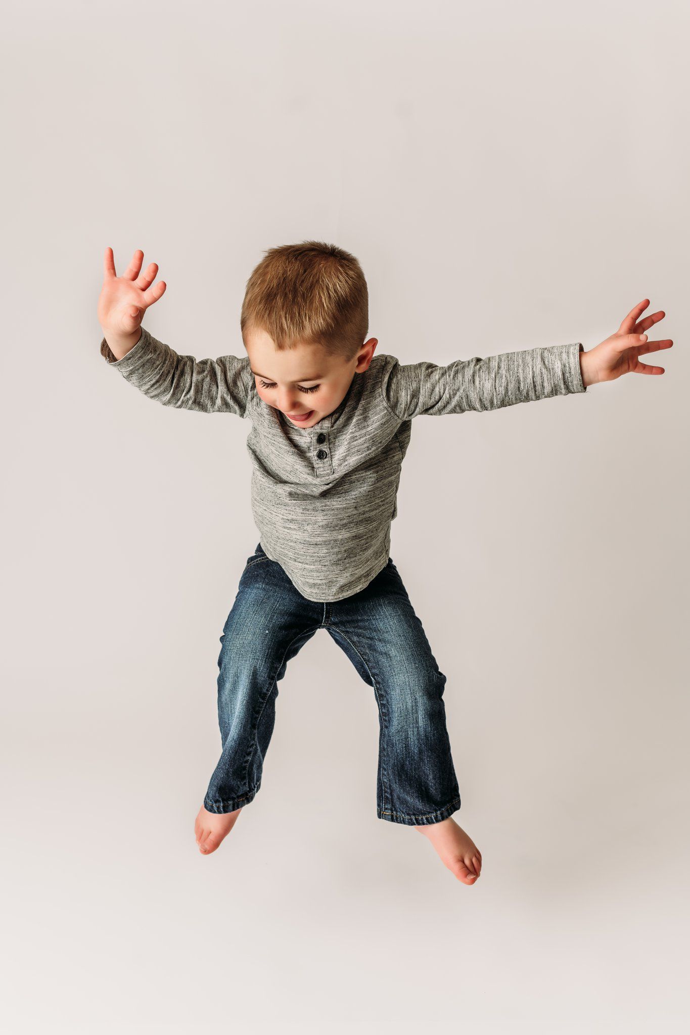 Child in gray shirt and jeans jumps with arms outstretched on white background.
