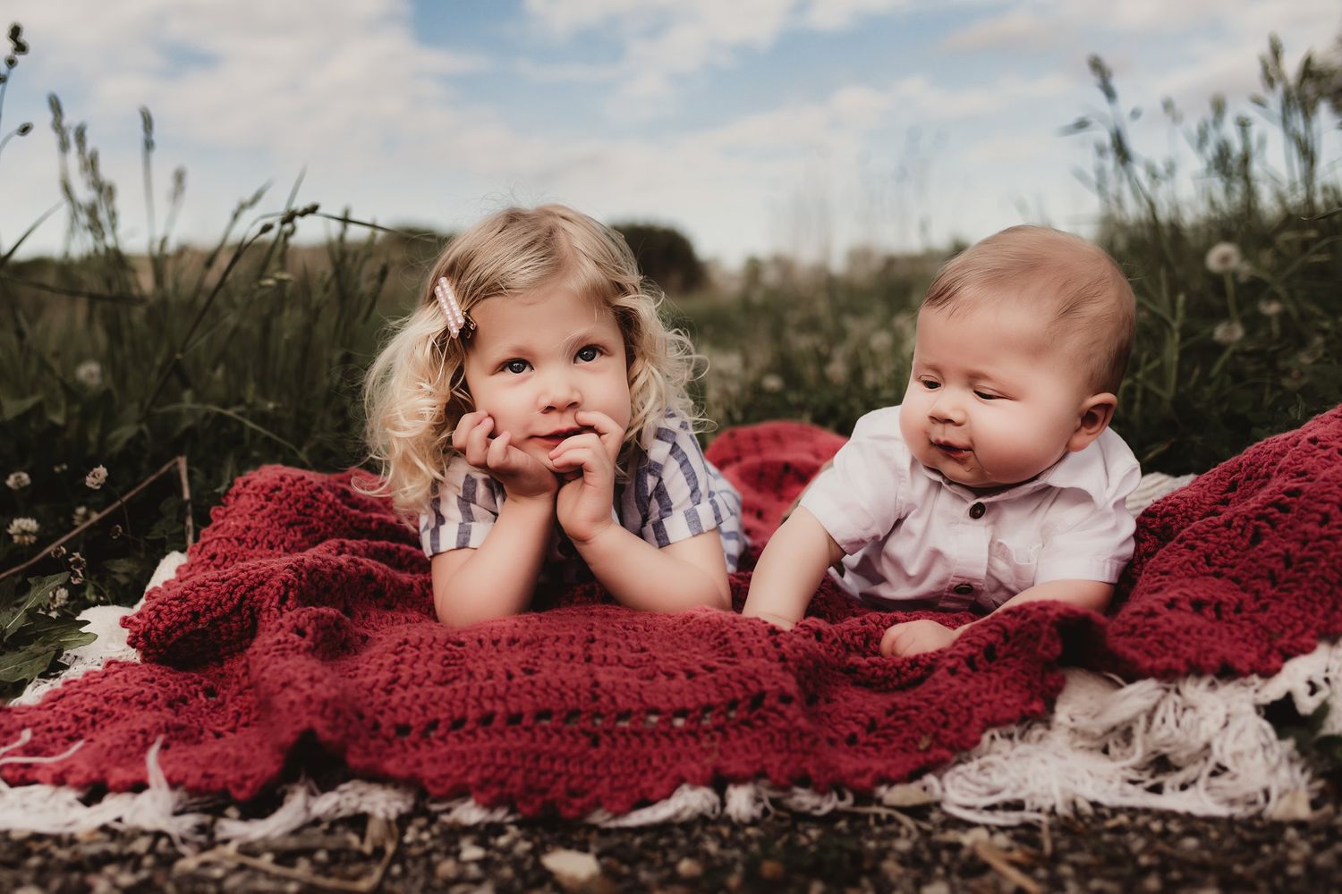 Candid portrait of two children lounging on a red blanket in a natural setting.