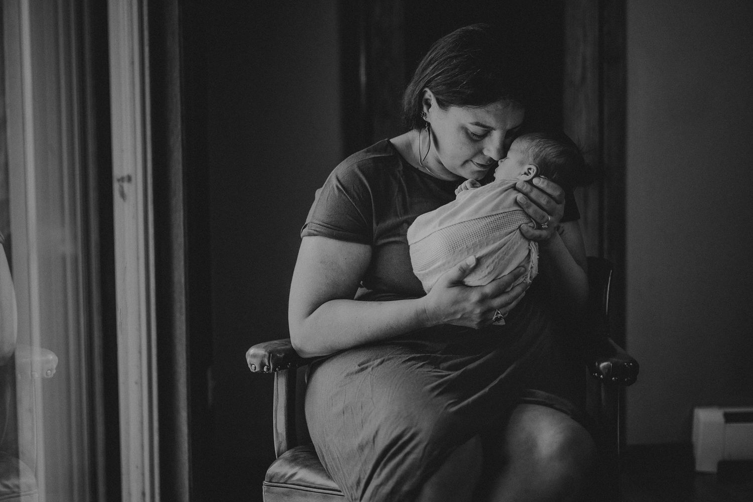 Parent sits in wooden chair holding and gazing lovingly at their swaddled infant.