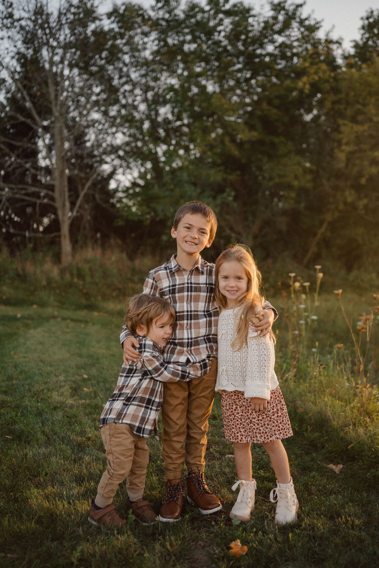Three children stand together outdoors at sunset wearing coordinated fall outfits in a grassy field.