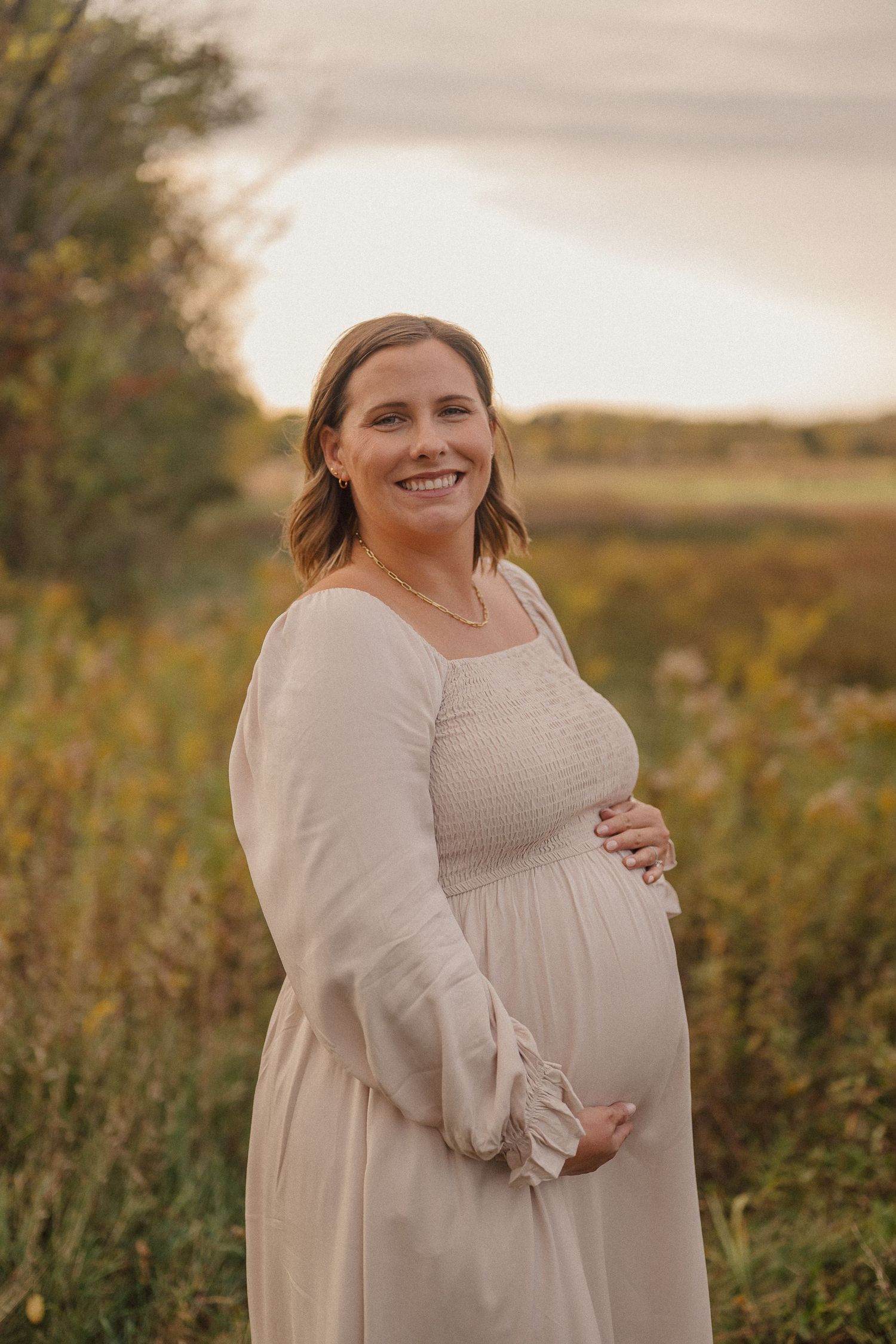 A pregnant person in a cream-colored dress poses in a golden field at sunset during a maternity photoshoot.