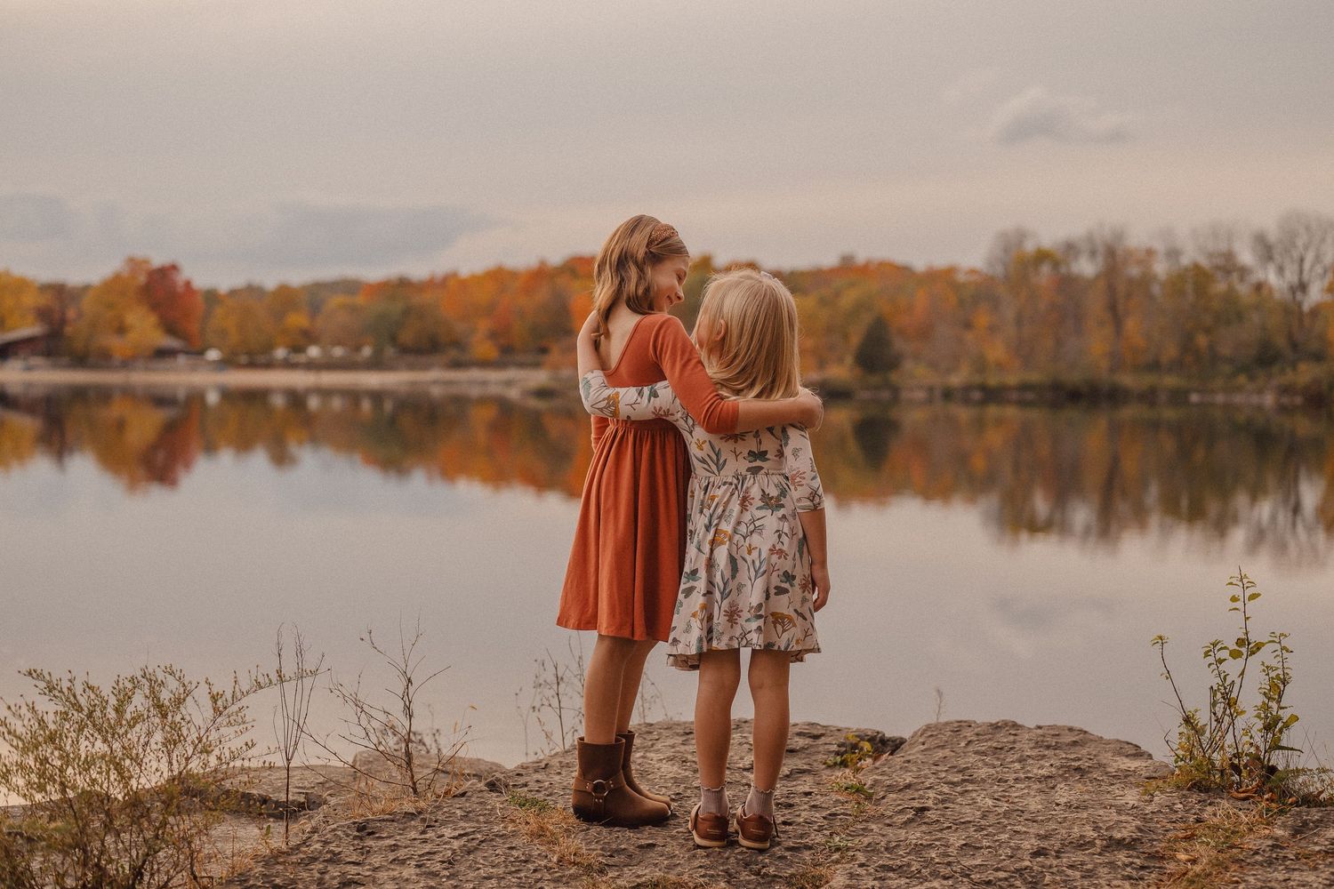 Two people stand at the edge of a tranquil lake during autumn sunset with fall foliage reflecting in the water.