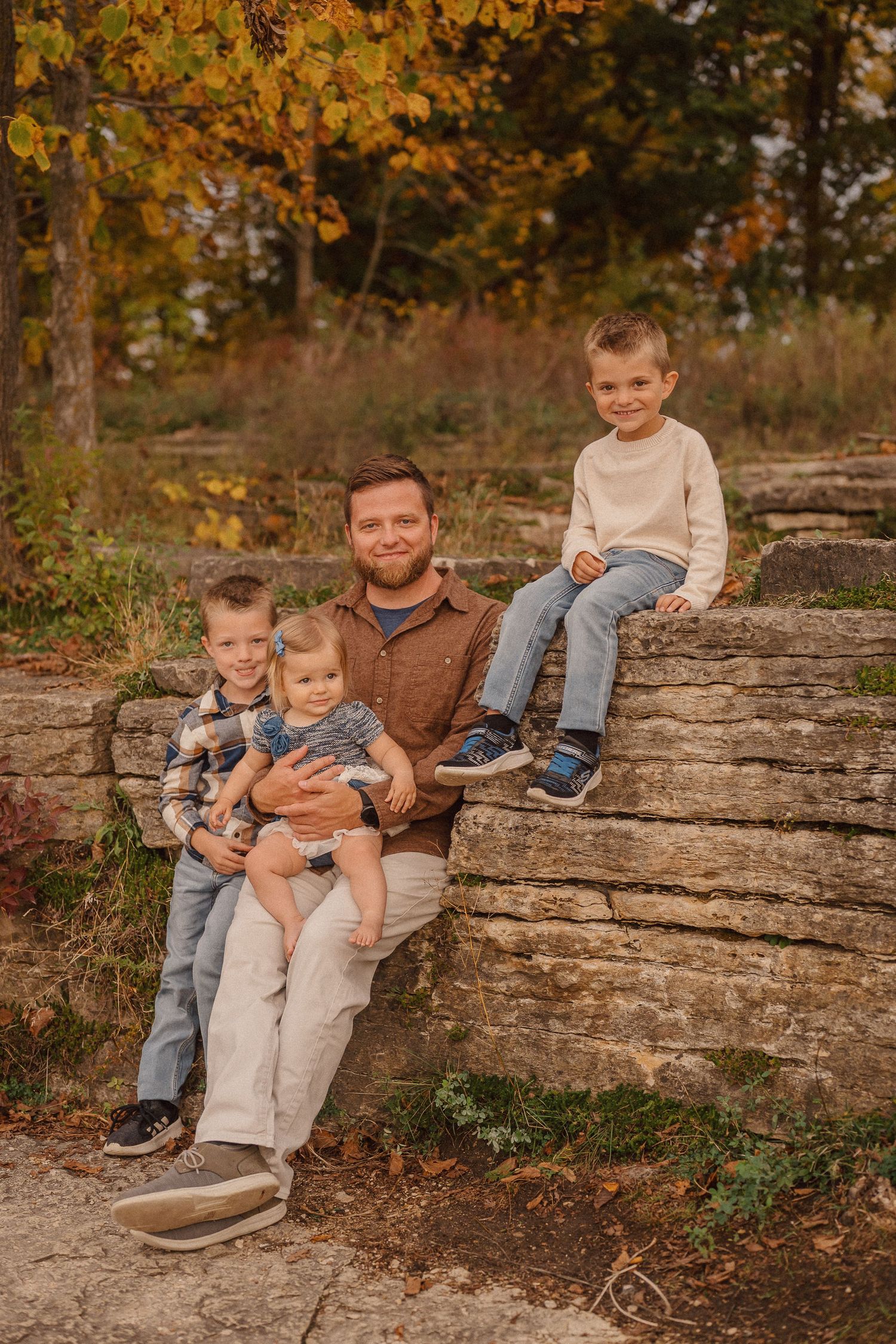 Family sitting together on a natural stone wall surrounded by autumn foliage in an outdoor setting.