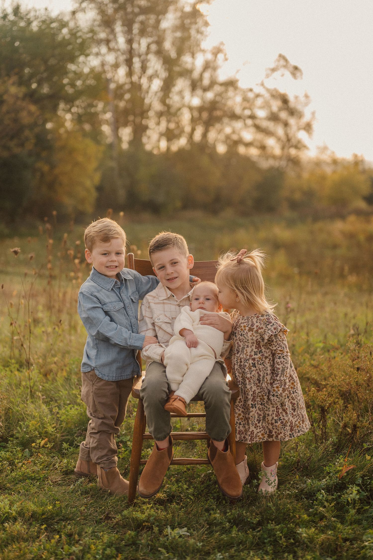 Three children share sweet moments while sitting together on a wooden chair in a sunlit autumn field.