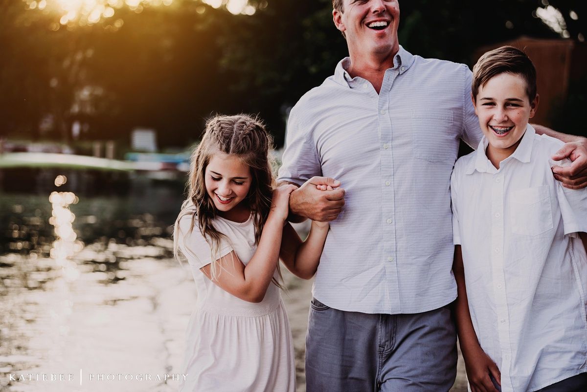 Three people share laughter and embrace by a lakeside marina at sunset.