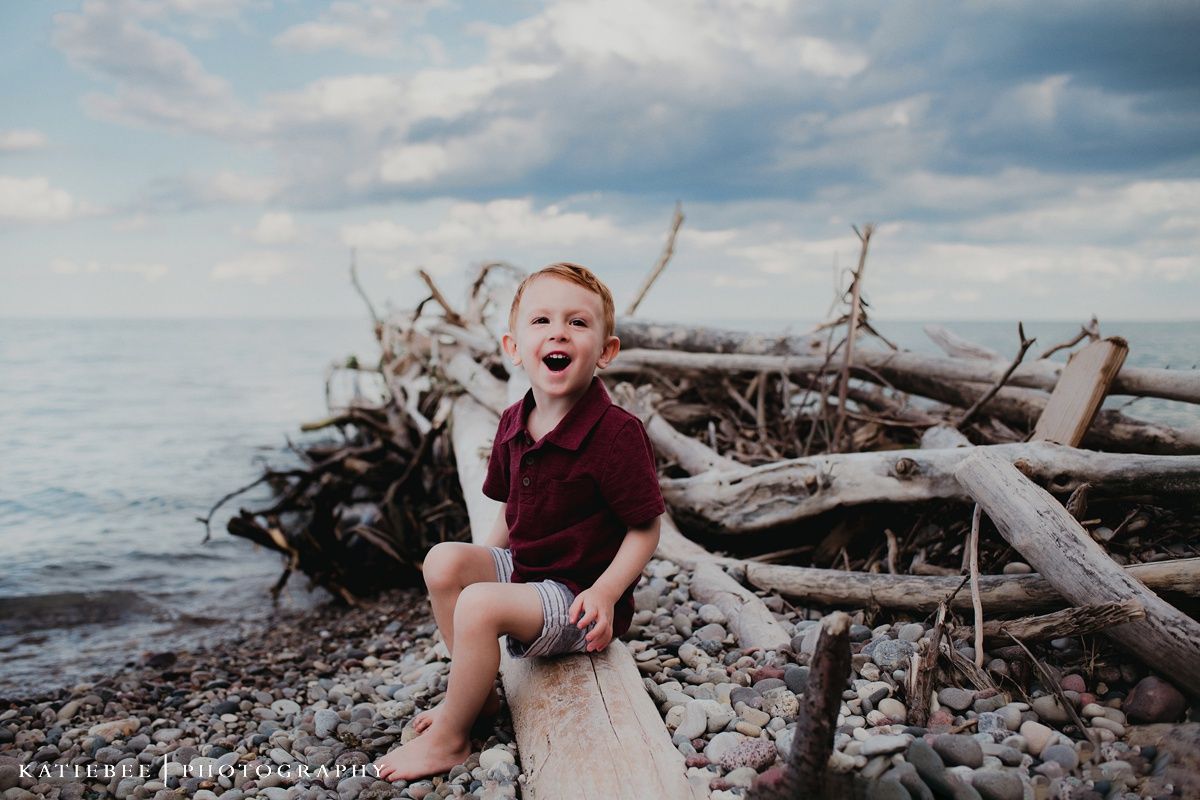 A child laughs while sitting on driftwood by the ocean on a cloudy day.