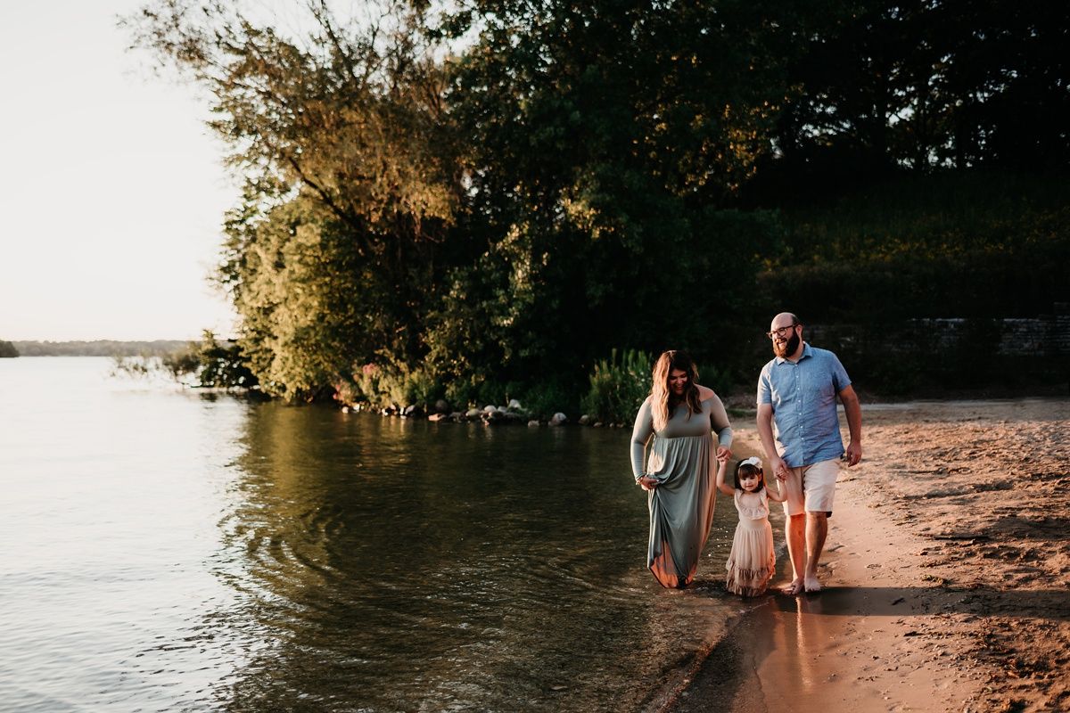 Family walks along peaceful shoreline at sunset with trees reflecting on calm water.