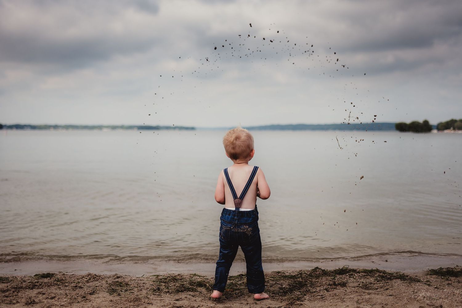 Small child in suspenders watching water drops arc through the air at a lakeside beach.