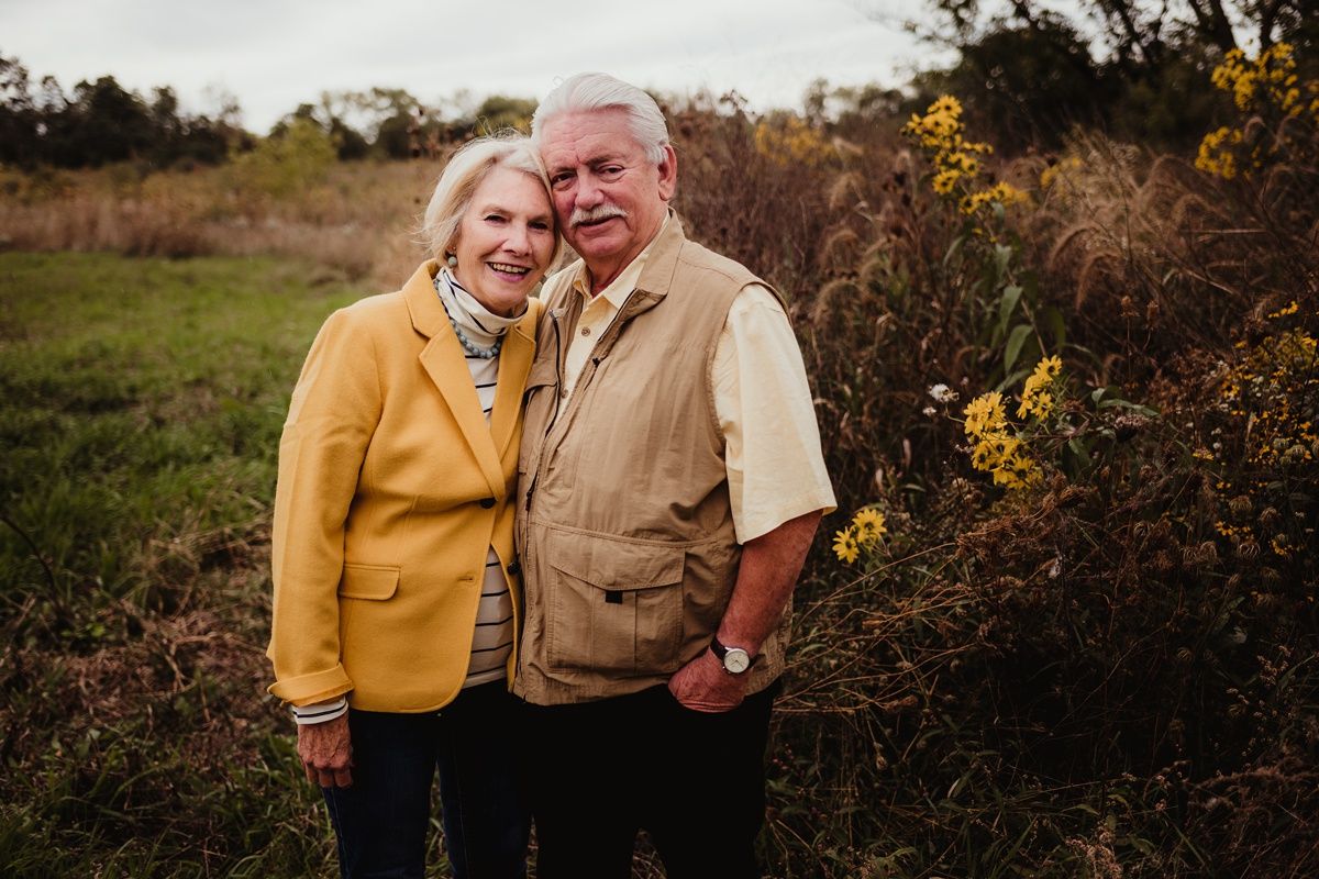 A couple wearing fall jackets poses together outdoors near autumn foliage.