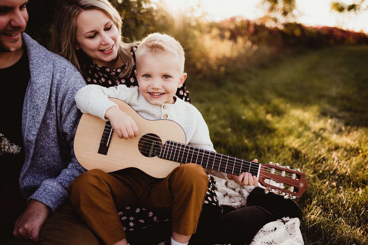 A child holds an acoustic guitar while sitting outdoors during golden hour sunset with others.