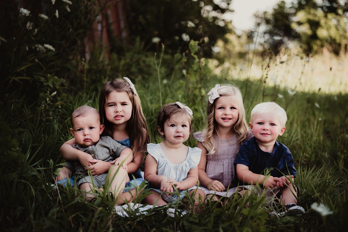 Five children sit together in a grassy field near an old barn structure.