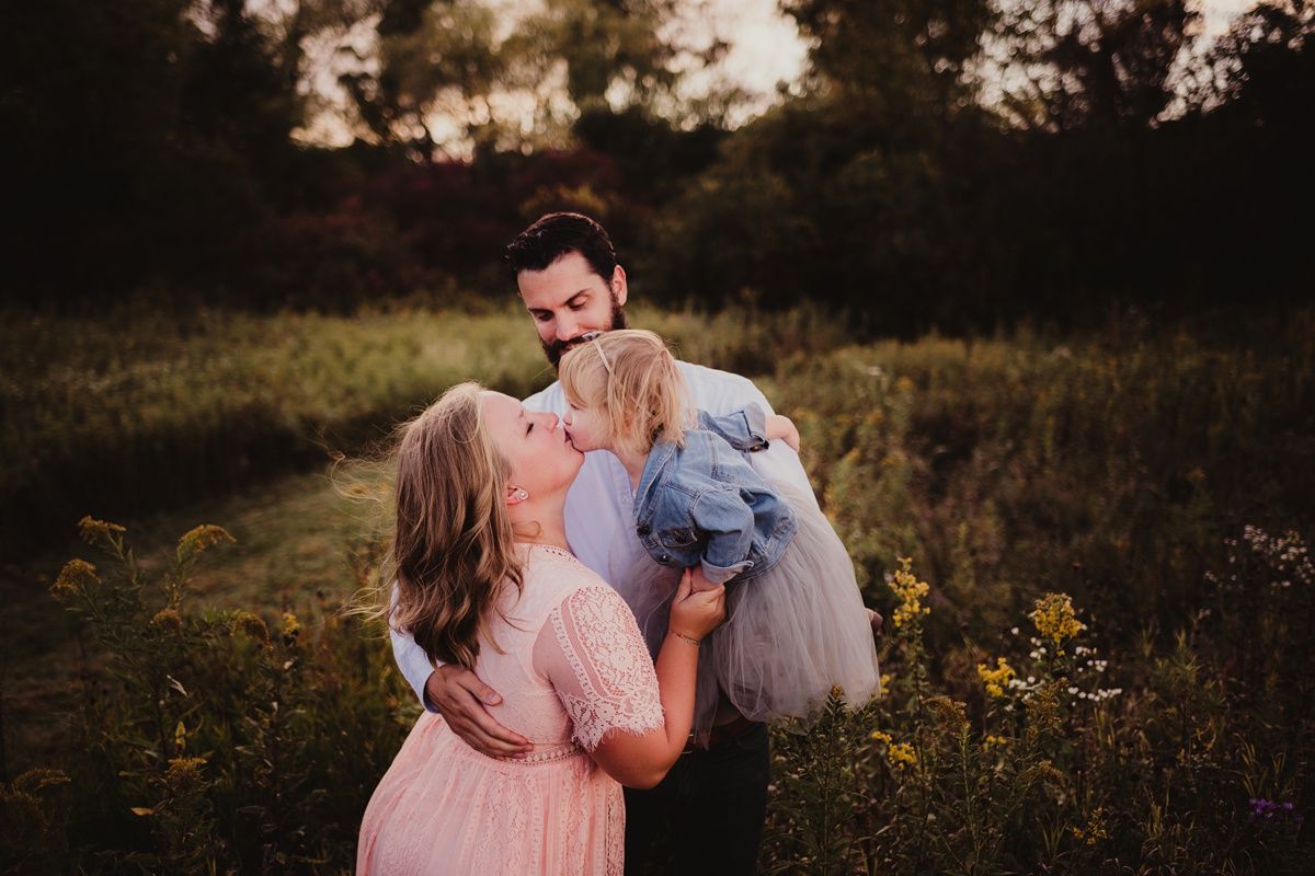 Family photo in a field at sunset with parent holding child while another parent looks on lovingly.