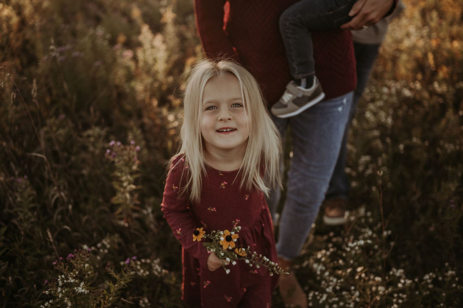 Child in burgundy dress playing in a wildflower field at golden hour.